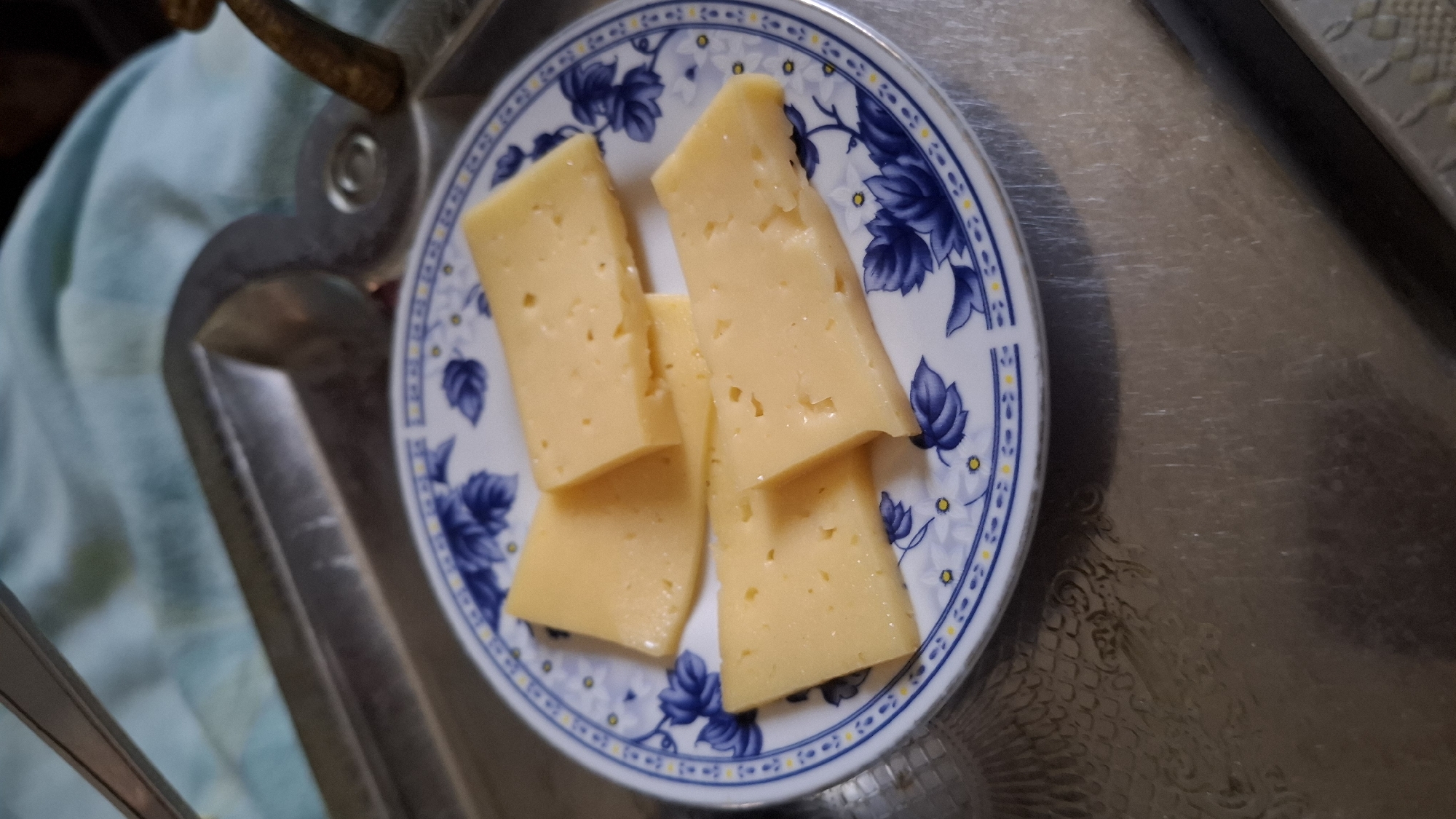 A close-up overhead shot shows four slices of yellow cheese arranged on a white plate with a blue floral pattern. The plate is resting on a silver serving tray with a textured surface. The tray is on top of a light blue fabric. The overall setting appears to be indoors, possibly in a kitchen or dining area, in the city of Shubarssu, Kazakhstan. There are no people visible in the image.