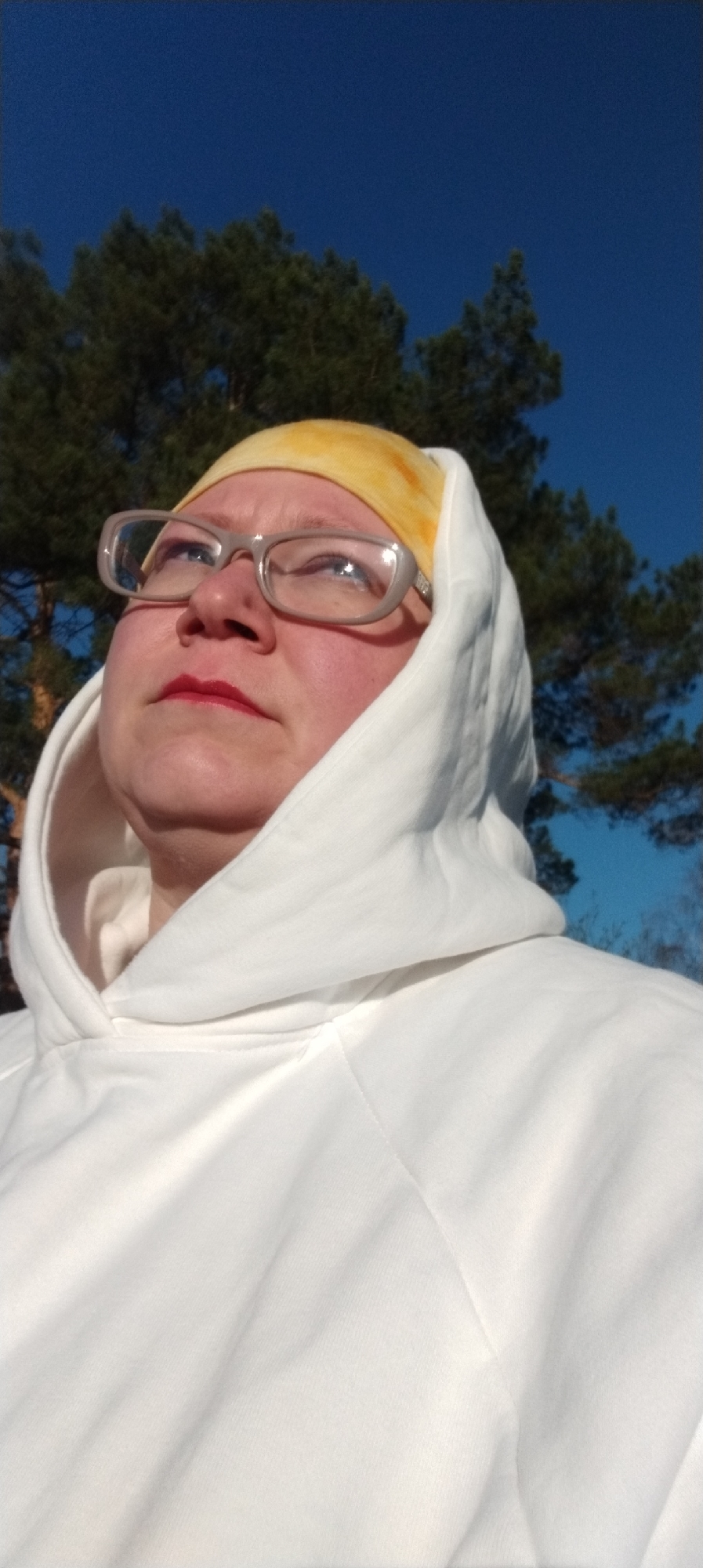 A woman with a yellow headscarf and glasses is wearing a white hoodie. She is looking up towards the sky. In the background, there are trees and a clear blue sky. The photo was taken in the city of Chaikovsky, Russia.