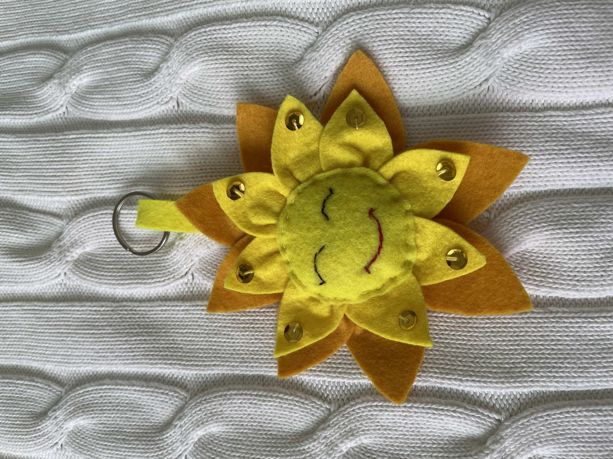 A top-down, eye-level shot shows a yellow and orange felt sunflower key chain with a smiling face on a white knitted blanket. The sunflower has a circular yellow face with black stitched eyes and a small red stitched smile. Gold sequins are attached to the yellow petals. The sunflower has two layers of petals, a yellow inner layer and an orange outer layer. The key chain is attached to the left side of the flower with a small loop of yellow felt. The blanket is a white cable knit pattern. The photograph was taken indoors in Лесной Пейзаж Кп (шарапово, Russia.