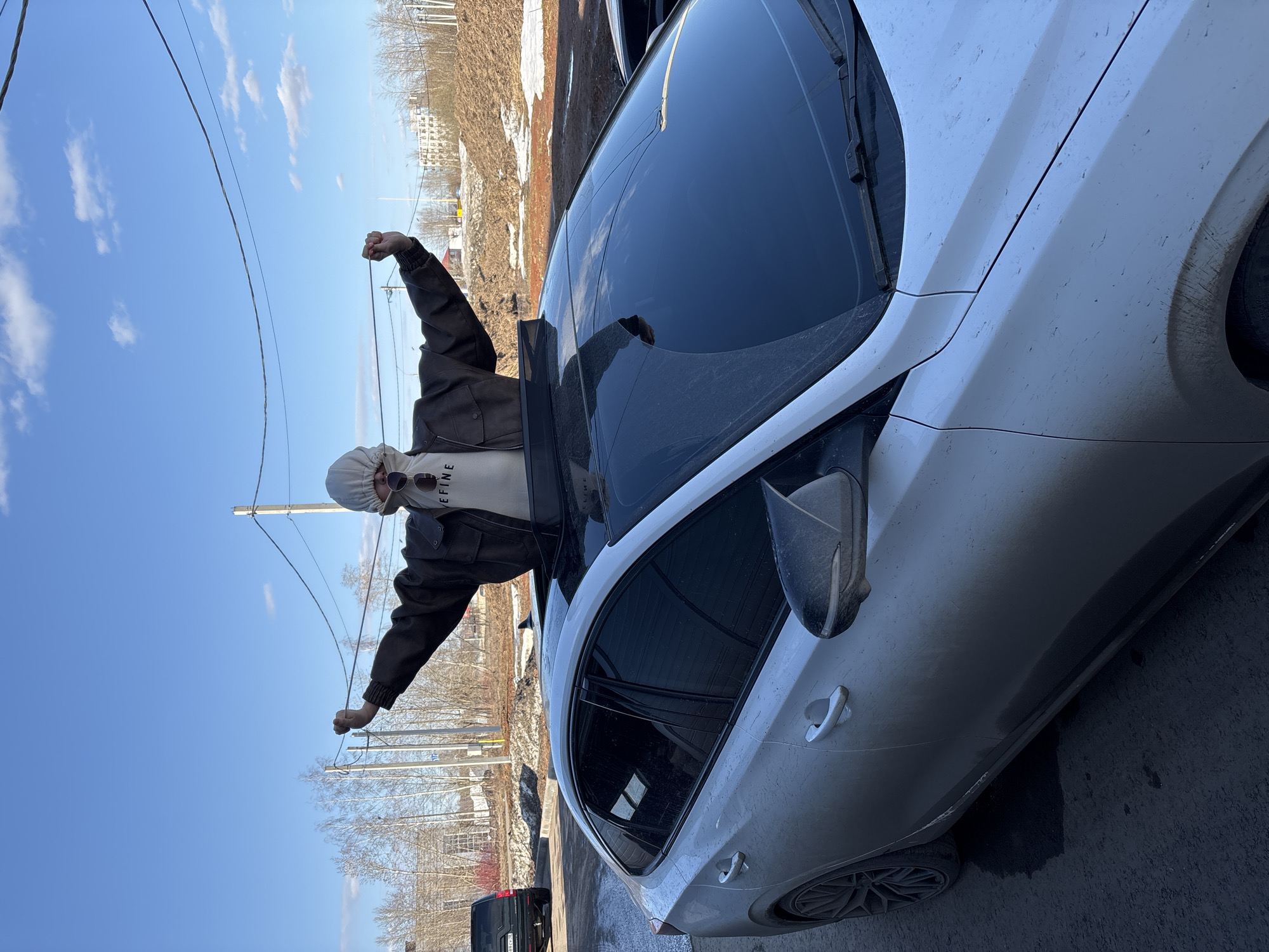 A person stands through the open sunroof of a dirty, white, modern car. The person is wearing a light-colored hoodie, a brown jacket, and sunglasses. The car is parked on a city street near a curb. Overhead power lines are visible, and in the background are trees, buildings, and patches of snow. The location appears to be in Izhevsk, Russia.