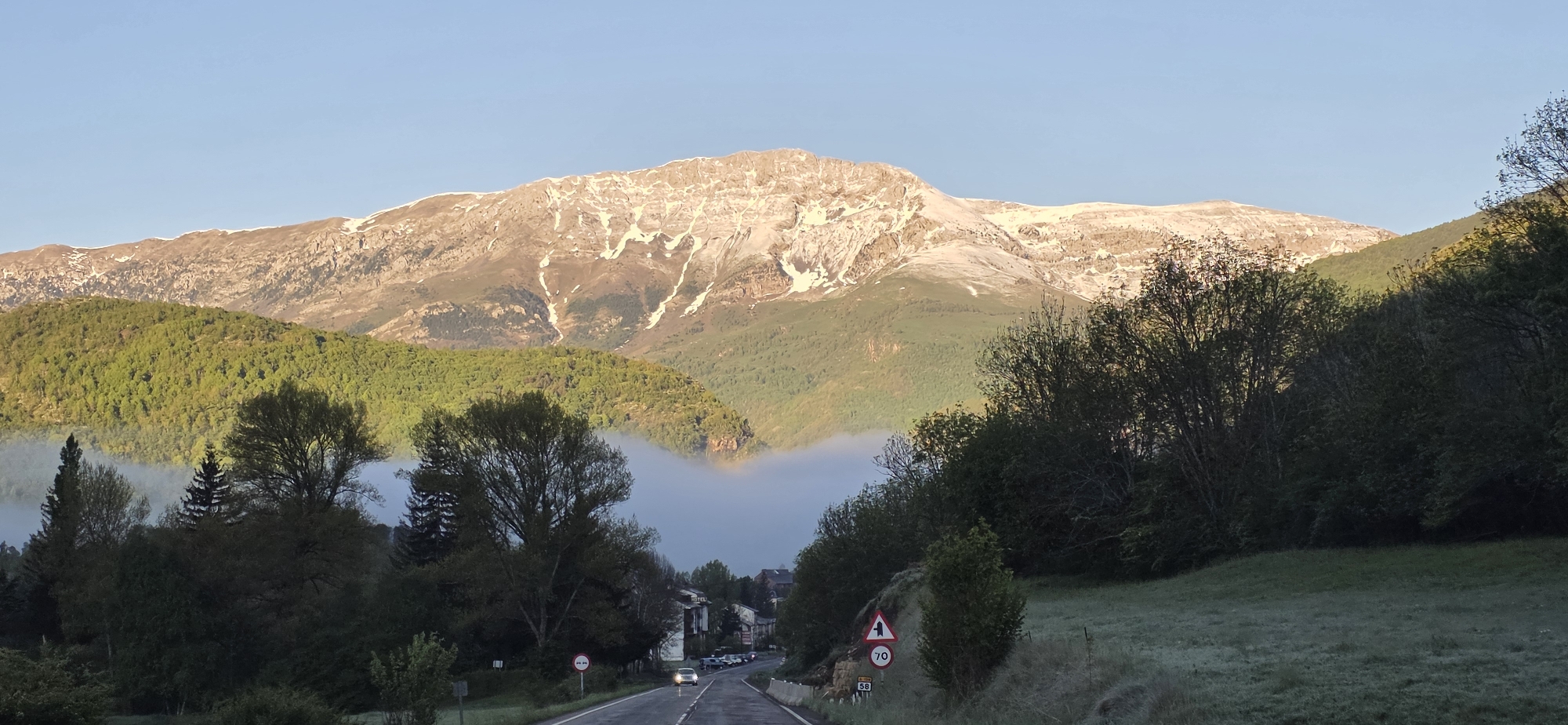 The image shows a scenic view in the city of Sahún, Spain. A road leads into the distance, flanked by trees and vegetation on both sides. A layer of fog partially obscures the view, creating a soft, misty effect. In the background, a large mountain range dominates the landscape, with snow-capped peaks visible. A road sign is visible on the side of the road. A few vehicles are seen on the road in the distance, suggesting some traffic activity.