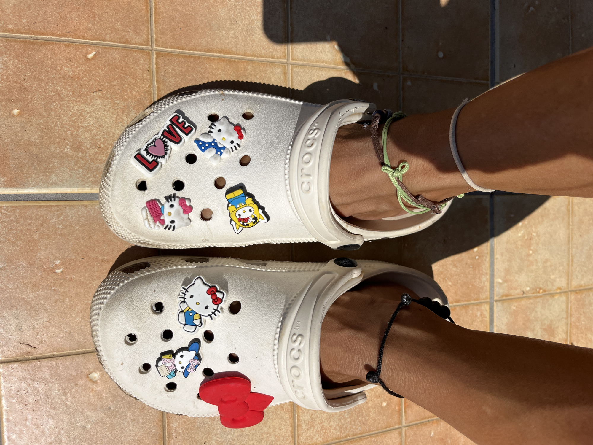 The image shows a pair of white Crocs on a person's feet. The left Croc has a large red bow charm and several Hello Kitty charms. The right Croc has the word "LOVE" with a heart, and other Hello Kitty charms. The Crocs are on a tiled surface. The person is wearing several bracelets on their ankles, including a black beaded one and green and brown string bracelets. The scene is outdoors, with shadows cast on the tiles. The location is specified as Phu Thuy Ward, Vietnam.