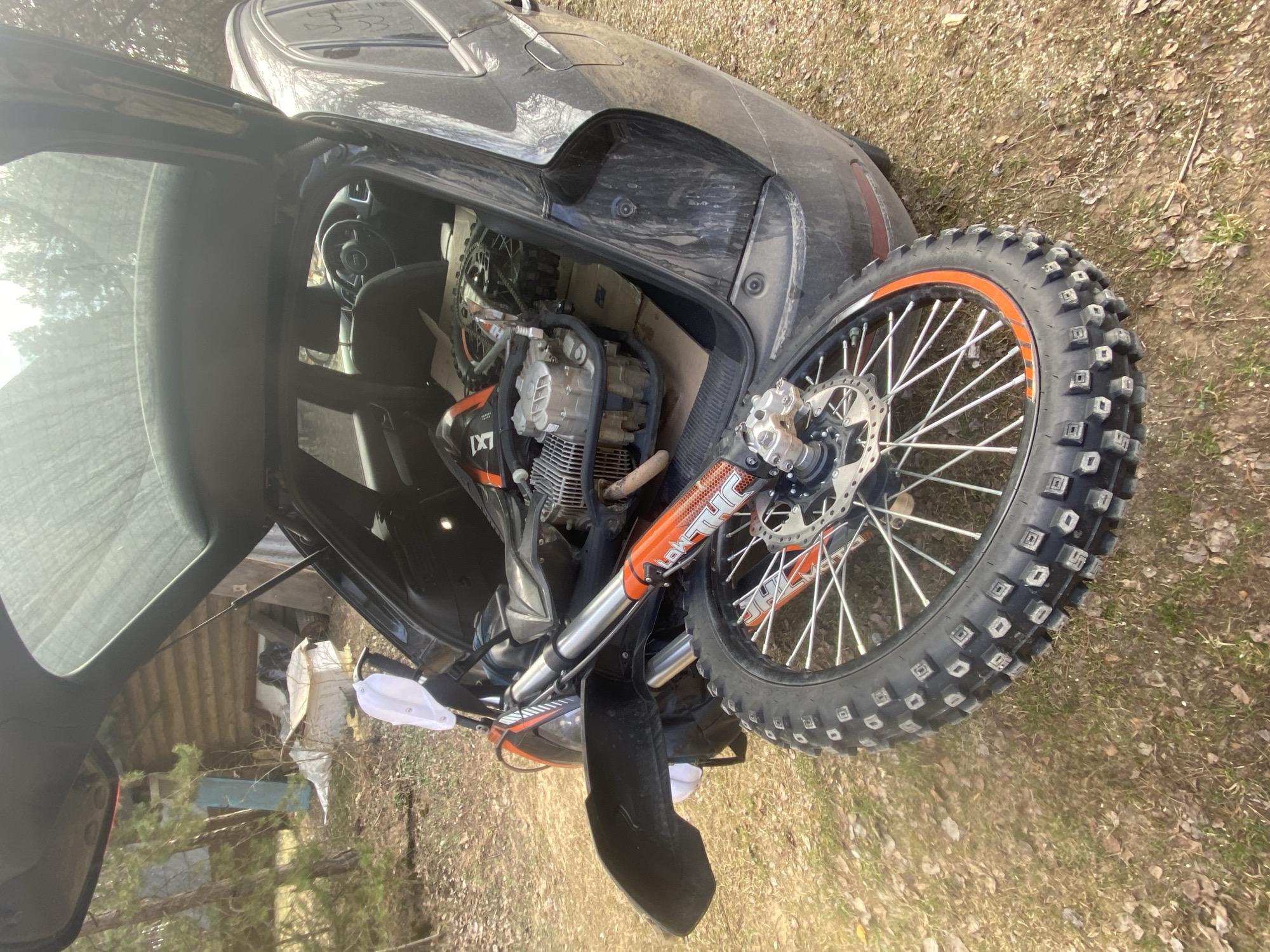 The image shows a dirt bike loaded into the back of a black Audi hatchback with the rear door open. The location appears to be outdoors, with the vehicle and bike sitting on dry grass with some leaves and twigs. The location is likely Парэчча, Belarus.