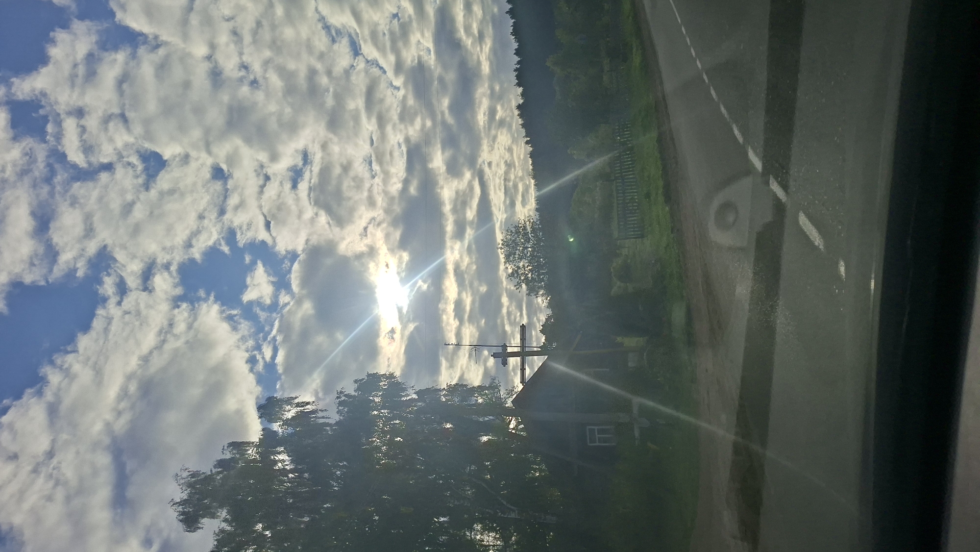 A vertical, slightly angled shot captures a rural landscape with a bright, partially clouded sky. The sun, positioned high and slightly off-center to the left, is the brightest point, emitting strong light rays that create lens flares across the image. In the mid-ground, a small, dark wooden structure with a window is visible. To the left of the structure, a tall, dark-barked tree with lush foliage dominates the scene. A cross, appearing to be made of wood and possibly with antenna elements, stands prominently near the structure. A light-colored wooden fence runs along the mid-right of the image, bordering a patch of grass. A paved road with white lines is in the foreground, angled towards the right side of the frame. The background is filled with a dense line of trees, forming a dark, forested horizon. The overall impression is that of a serene, somewhat overcast day in a rural setting, possibly in the Сітцаўскі сельскі Савет, Belarus.