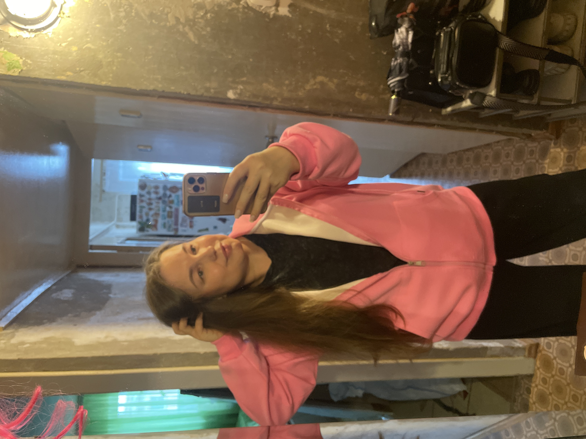 The image displays a mirror selfie of a young woman taken indoors, rotated 90 degrees clockwise. The woman has long, dark brown hair and is wearing a pink zip-up hoodie over a black top and black pants. She holds a gold or beige-cased smartphone, resembling an iPhone, with her right hand to capture the reflection. Her expression is neutral, with a slight smile, as she looks towards the phone's camera.
The reflection in the mirror reveals a room with distressed and discolored walls or ceiling, suggesting an older space or one undergoing renovation. An illuminated circular overhead light fixture is prominently visible in the upper left of the reflection. Other reflected objects include a dark bag or backpack in the upper right quadrant. The lower portion of the reflection shows patterned tiles or wallpaper, along with a glimpse of bright light and patterned items through an opening in the background, possibly a doorway or window. A small section of pink, resembling yarn or string, is visible in the bottom left. The scene is lit by artificial light. No discernible text is visible within the image. The location context for this scene is Izhevsk, Russia.