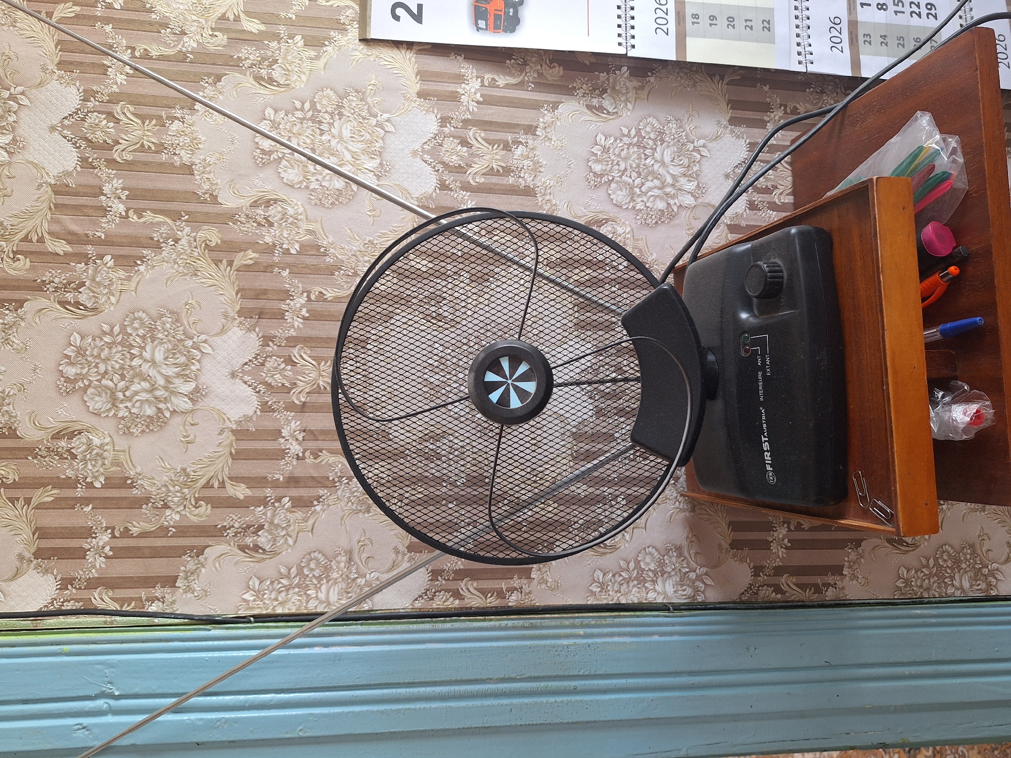 The image shows a close-up of an indoor television antenna with a black circular mesh dish, mounted on a black base. The antenna is positioned on a wooden shelf. To the left of the antenna, a portion of a light blue ribbed surface is visible, possibly a door or wall panel. The background is wallpaper with a floral pattern featuring cream and brown tones. To the right of the antenna, a portion of a calendar for the year 2026 is visible, displaying numbers and some text in Russian. The calendar appears to be a desk calendar with a spiral binding. A few items are on the wooden shelf below the antenna, including paperclips, a plastic bag containing colored pencils or pens, and the tip of a blue pen and orange pencil. The overall scene suggests an indoor setting, possibly a living room or study in a residential area within Блеўчыцкі сельскі Савет, Belarus.