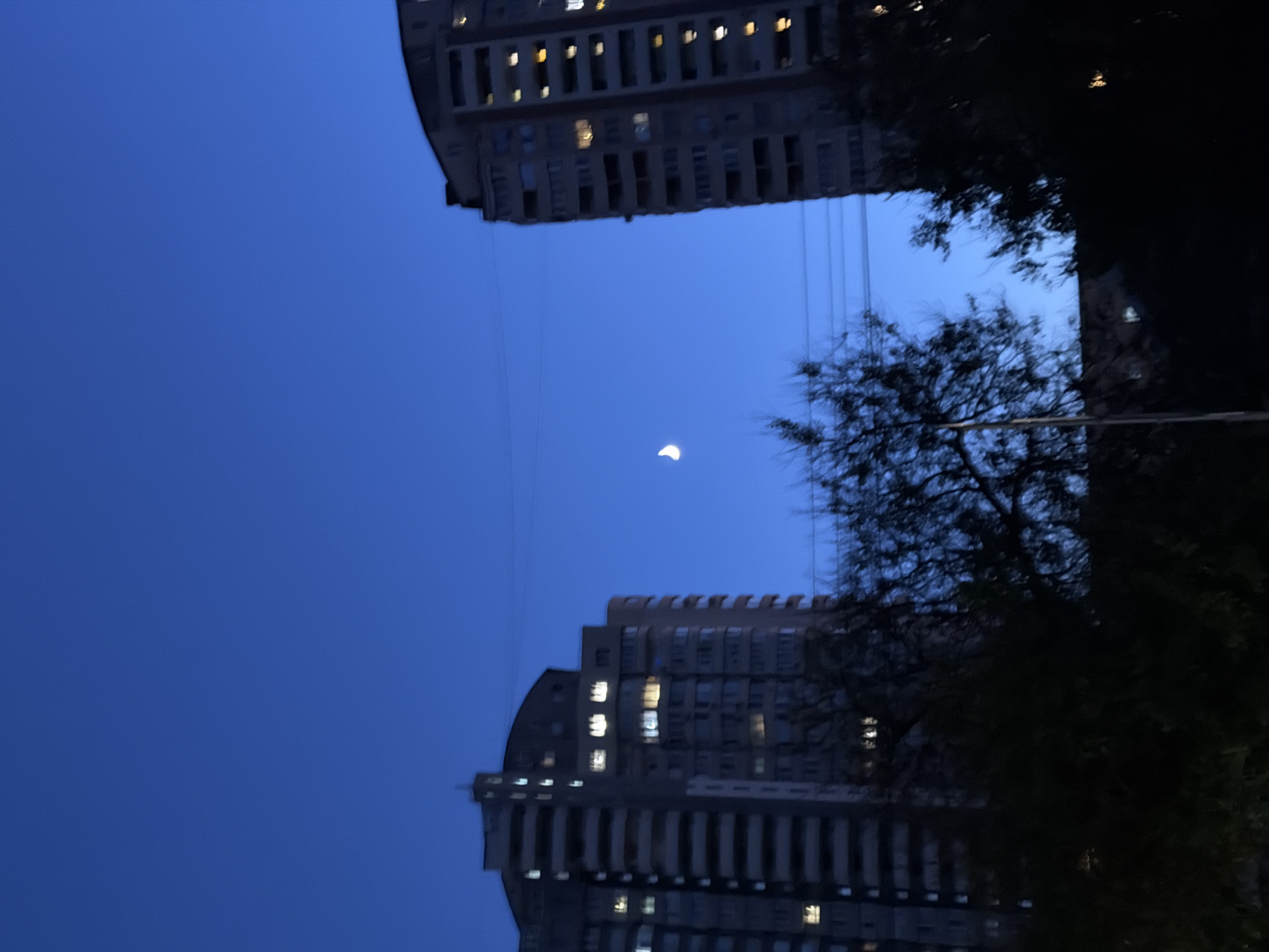 The image depicts a night sky over Kyiv, Ukraine, with two tall apartment buildings visible. The sky is a deep blue, indicating it is either dusk or dawn. A crescent moon is visible in the sky between the two buildings. The apartment buildings have many windows, some of which are illuminated with warm light, suggesting people are home. Silhouetted trees are in the foreground, obscuring the lower parts of the buildings and the ground. Electrical wires are also visible stretched between the buildings.