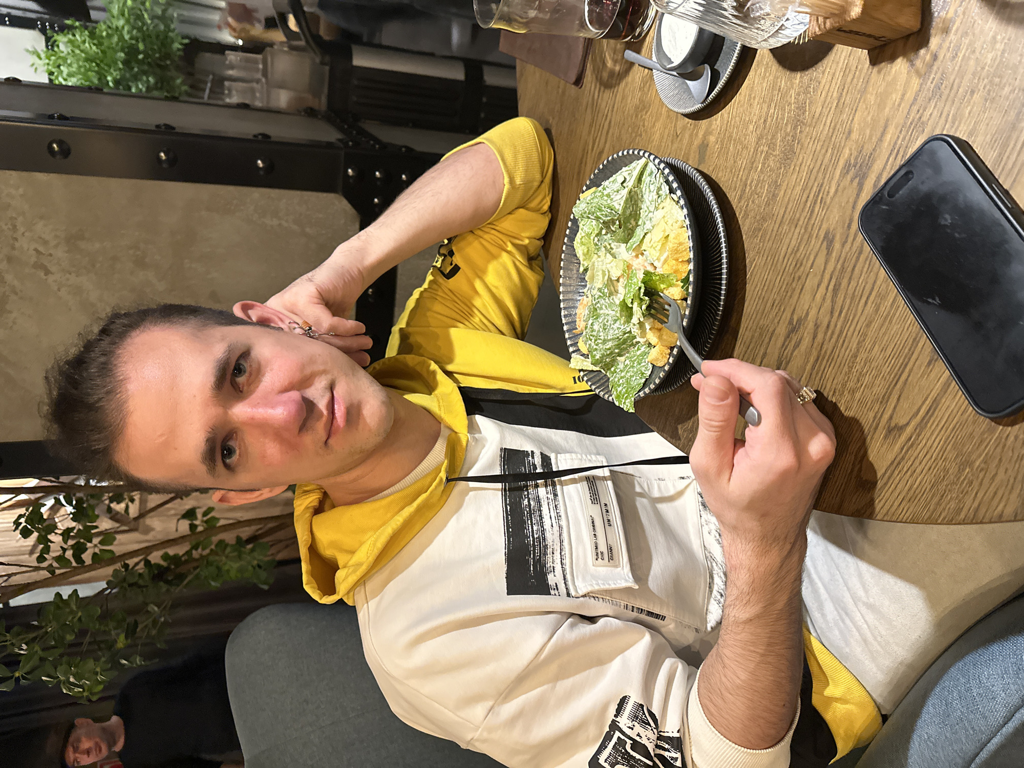 A man is seated at a wooden table in what appears to be a restaurant or cafe. He is wearing a white and yellow hooded sweatshirt, and he has a salad on a black plate in front of him, which he is about to eat with a fork. A black smartphone is on the table beside the plate. Other items on the table include glassware and condiments. The background shows parts of the restaurant interior, including plants and metal structures. While the location in Saratov, Russia, is mentioned, there are no visual indicators in the image to confirm the specific city or country.
