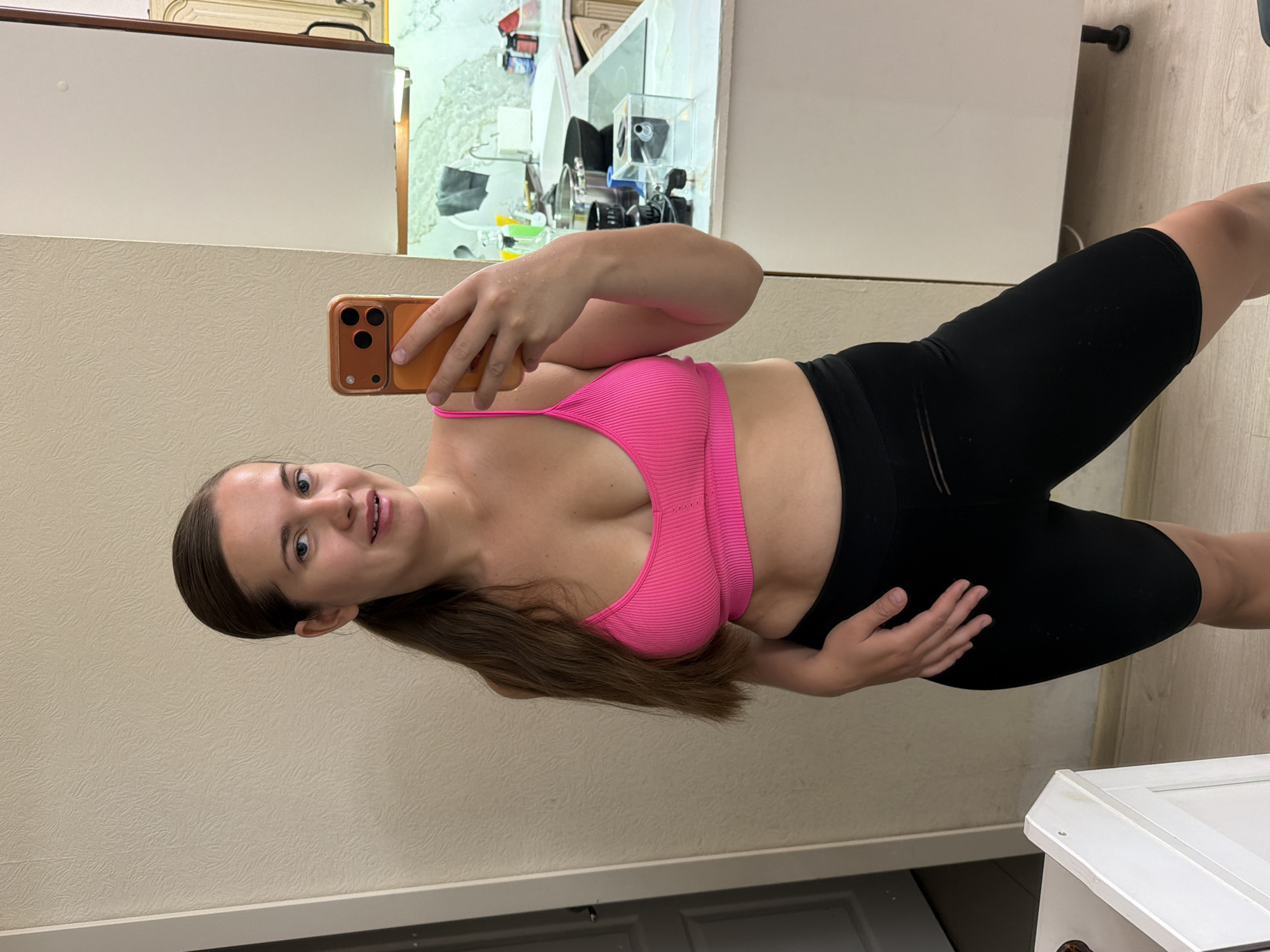 A female, appearing in her 20s or 30s with long brown hair styled in a low ponytail, captures a mirror selfie in an apartment located in Sochi, Russia. She is clad in a bright pink ribbed sports bra with thin straps and black mid-thigh length cycling shorts. Her right hand, holding an orange-cased smartphone with a multi-lens camera, is visible. Her left hand rests near her hip. She displays a slight smile with parted lips, and a small hoop earring is visible in her left ear. The background features a light beige textured wallpaper. To the left, a white interior door with a dark handle is partially visible, alongside a white cabinet. To the right, a kitchen area is present, equipped with white upper and lower cabinets, a light-colored countertop, a stainless steel sink with a faucet, and a black induction cooktop. Various kitchen items, including bottles, a frying pan in the sink, a grey towel, and a clear rectangular container, are arranged on the counter. The flooring is a light wood-look laminate or tile. Under-cabinet lighting illuminates the counter.