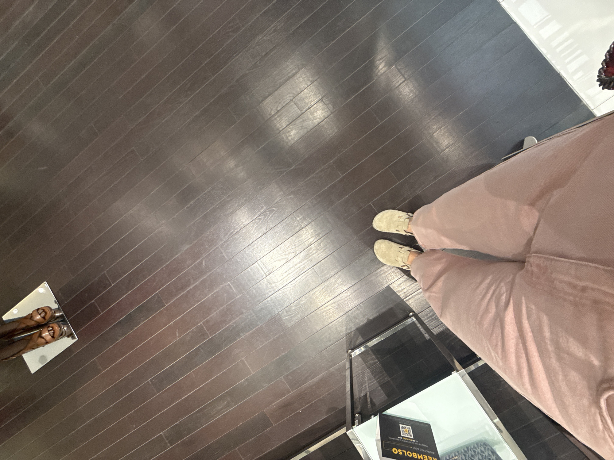 A photograph taken at floor level shows a dark wood floor with a person wearing light brown shoes and pink pants. To the left is a display stand, and in the lower right is a glass shelf holding a product labelled "REEMBOLSO." The location is likely a retail store in Puebla, Mexico.