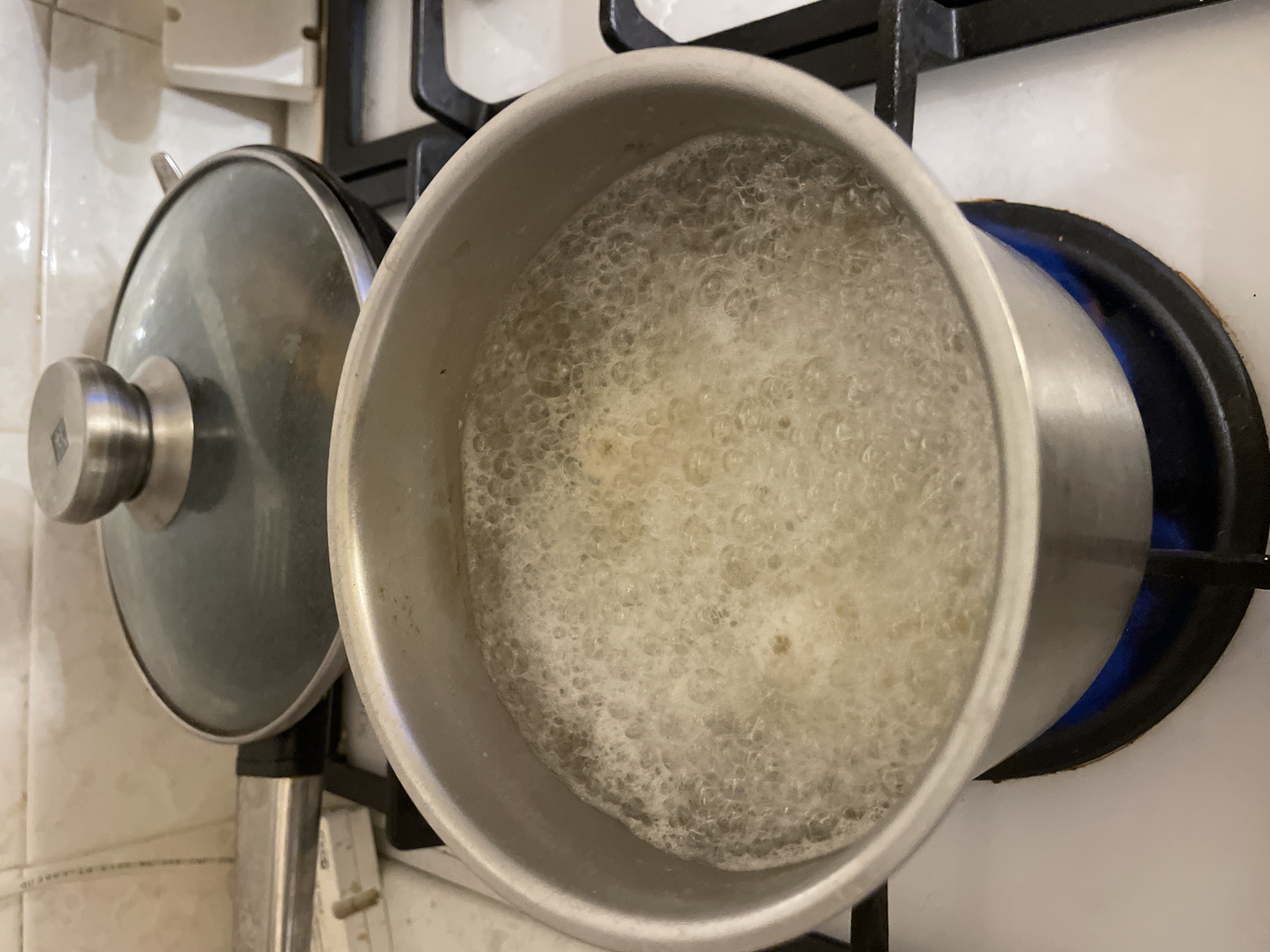 A top-down, close-up shot displays a metal saucepan filled with boiling liquid on a black stovetop burner. A metal lid is positioned to the side of the saucepan. The tiles of the kitchen wall are visible in the background. No people are visible in the image. The location is unknown, however, it could be the city of Moscow, Russia.