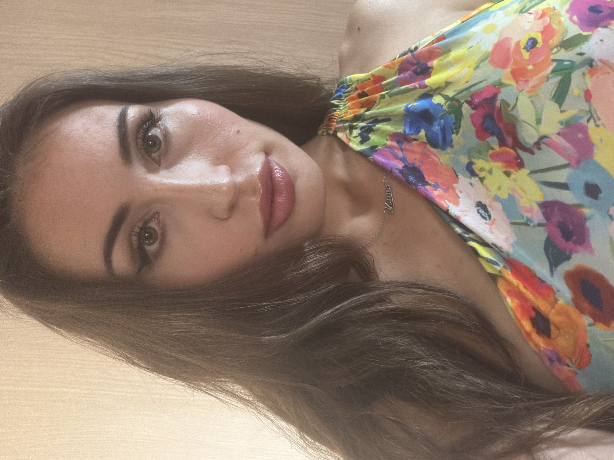 The image is a close-up portrait of a woman with long, dark brown hair. She is wearing a colorful floral-print dress with a halter neckline. A delicate silver necklace with the name "Lana" is visible around her neck. The background appears to be a plain, light-colored wall. The lighting suggests an indoor setting. There is no indication of the location being the city of Tersefanou, Cyprus.