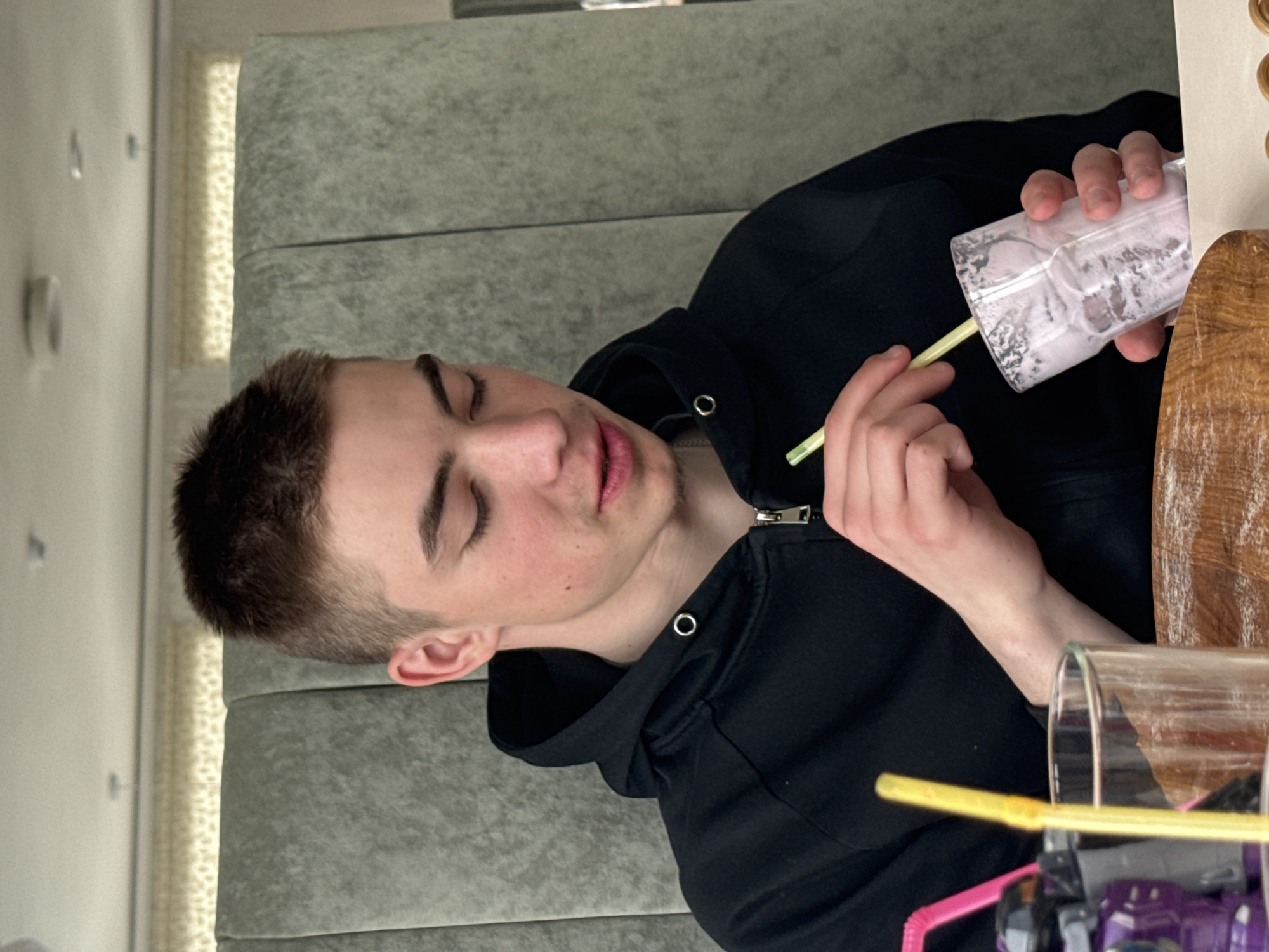 A young man with short brown hair is seated and appears to be looking down at a table. He is wearing a black hooded sweatshirt. He is holding a glass filled with a light pink beverage and a yellow straw. The location may be a café or dining area in Chaikovsky, Russia, based on contextual clues. Other straws and potentially a toy are visible on the table near another glass.