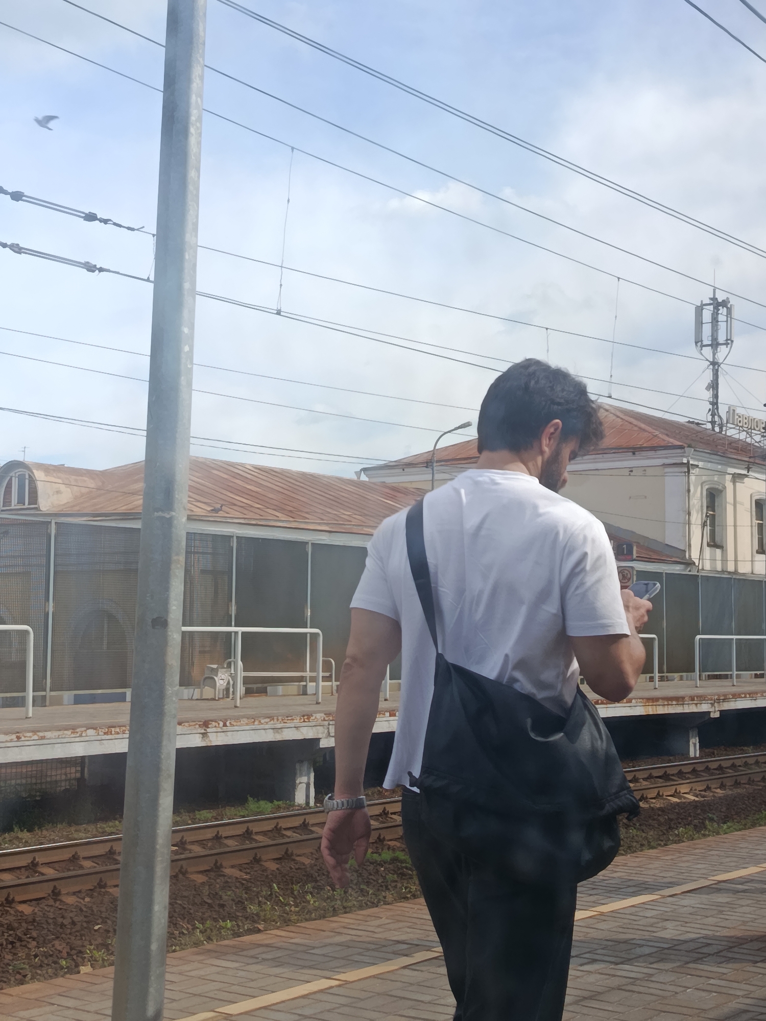 A man with dark, curly hair is standing on a train platform in Pavlovsky Posad, Russia, looking down at his phone. He is wearing a white t-shirt and dark pants, with a black messenger bag slung over his right shoulder. A silver watch is visible on his left wrist. In the background, an old, rust-colored train station building with arched windows stands behind a metal fence. The platform is paved with bricks, and railway tracks run alongside it. Overhead, power lines crisscross the sky, and a transmission tower is visible in the distance. A bird is flying in the upper left corner of the frame.