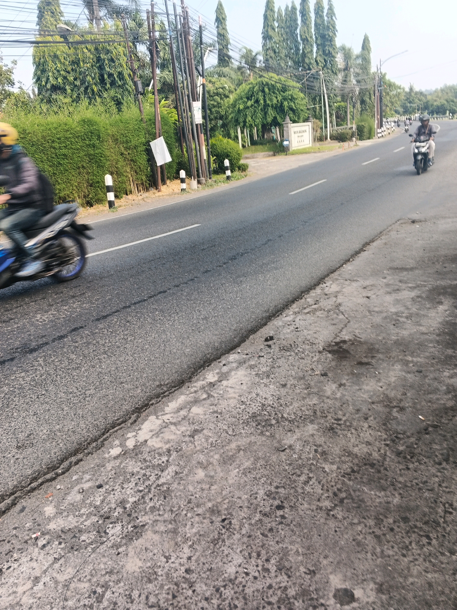 This is a photograph taken in Ungaran, Indonesia, showing a road with a motorcycle rider. The road is paved and appears to be a two-lane road with a white line marking the lanes. The rider is wearing a yellow helmet and dark clothing, and is on a blue and white motorcycle. The motorcycle is in motion, indicated by the blur of the wheels.
To the left of the road, there is a dense hedge and a series of utility poles with many wires. There are also some white and black striped bollards along the side of the road. Further in the background, there are many tall, slender trees and some smaller green bushes. A sign that reads "Multi Bakor" is visible on a pillar near the road.
To the right of the road, another motorcycle rider is visible in the distance. The pavement on the right side of the road appears to be rougher and more worn than the road itself, with some scattered debris. The sky is visible in the background and appears to be clear. The overall lighting suggests it is daytime, likely in the morning or late afternoon.