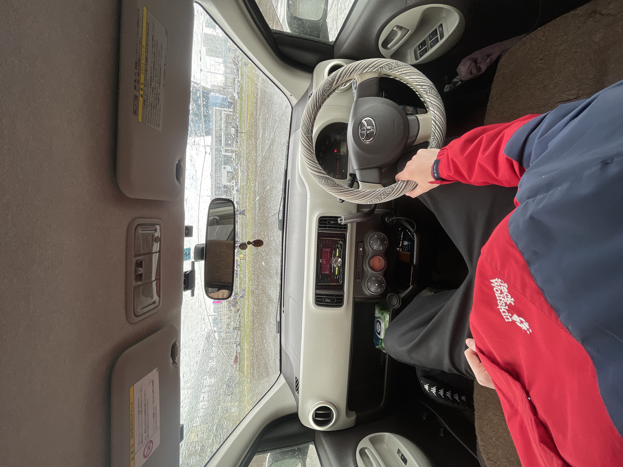 The interior of a car is shown in the image. The car is a Toyota, as evidenced by the logo on the steering wheel. A person is sitting in the driver's seat, holding the steering wheel. The person is wearing a red and blue jacket with the logo "Jack Wolfskin". Rain is visible on the windshield, obscuring the view outside. The car's dashboard is a light color. Based on the writing in the image, the location is Krasnoyarsk, Russia.