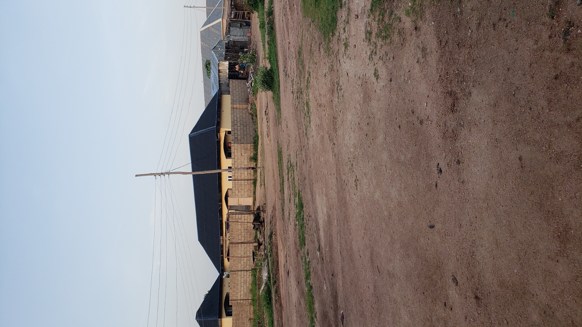 In Jauro Yinu, Nigeria, a street view captures a residential area characterized by a dirt road in the foreground. Beyond the road, a low-lying brick wall encloses several structures. The main building visible is a light yellow house with a dark, sloped roof, featuring arched windows and doorways. Adjacent to this house, and partially obscured, is another similar structure also with a dark, sloped roof. To the right, corrugated metal structures with sloped roofs are present, suggesting smaller dwellings or workshops. A wooden utility pole with multiple wires stands prominently in the middle ground, near the main house. The environment appears to be dry, with sparse patches of green vegetation and scattered debris on the dirt road. The sky is a pale blue, indicating either early morning or late afternoon light. No people are clearly visible in the scene.