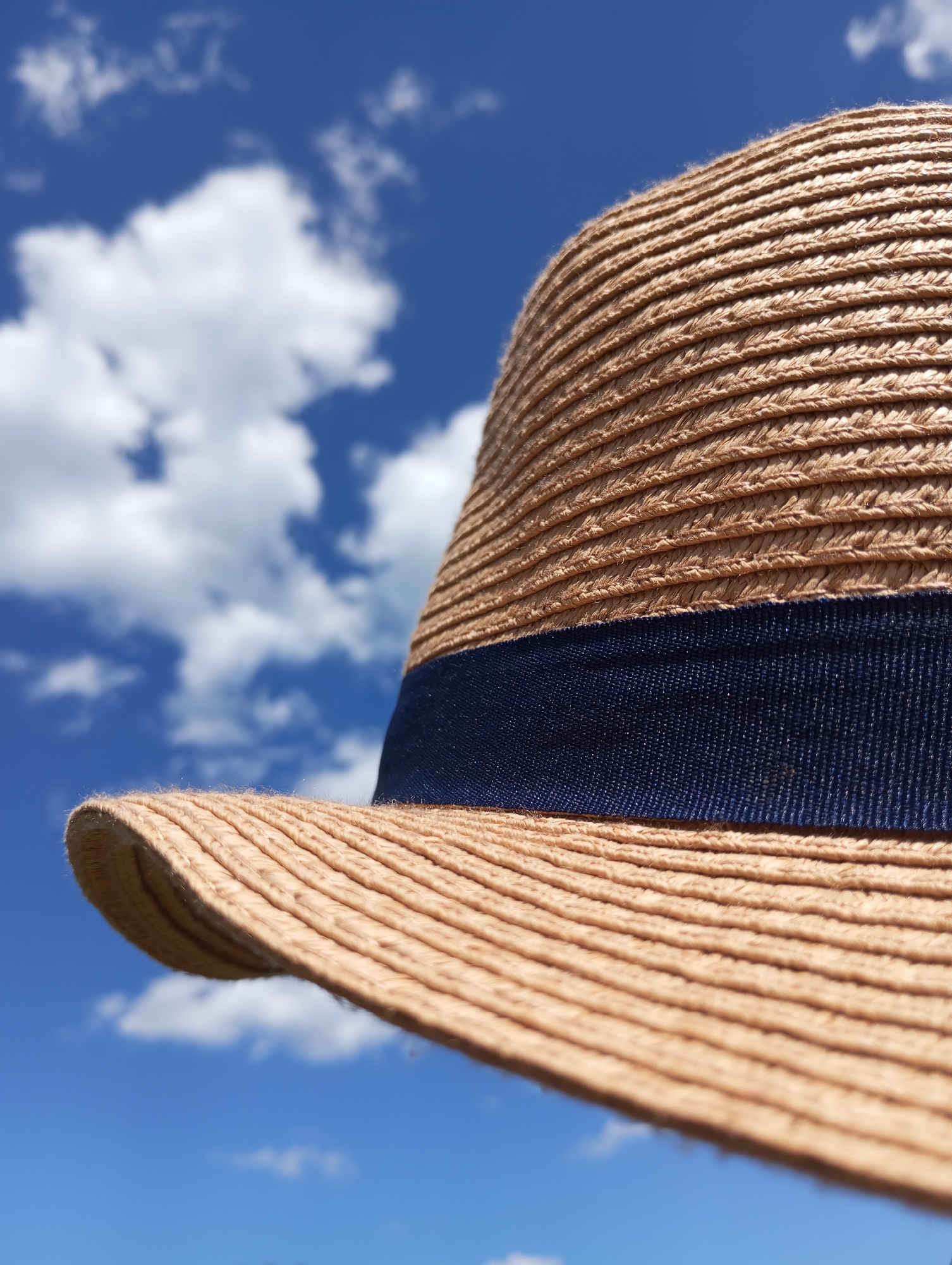 A close-up shot of a straw hat with a dark blue ribbon around its base, set against a backdrop of a bright blue sky dotted with fluffy white clouds. The brim of the hat is in the lower left corner of the frame. The city of Буренка, Russia is not present in the image.