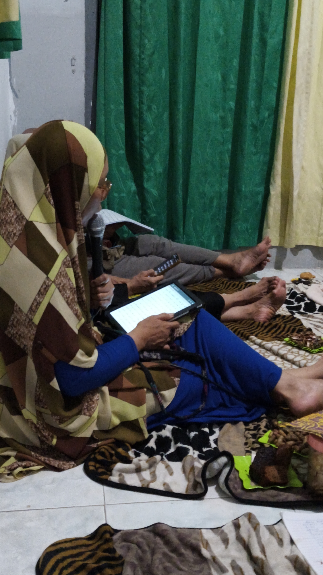 Two individuals are seated on the floor of a room. The room appears to be in Dalegan, Indonesia, based on the context provided. The floor is tiled and covered in various patterned blankets, including tiger stripes, cow print, and abstract designs. Behind the individuals are green and yellow curtains.
The person in the foreground, wearing a blue long-sleeved shirt and blue pants, is holding a tablet. They are also wearing a hijab with a geometric pattern of brown, beige, and yellow. A microphone is held in their left hand.
The second individual is seated behind and to the right of the first. They are wearing grey pants and a grey top. This person is holding a remote control in their right hand and a book or papers in their left hand. The feet of both individuals are visible and appear bare.
On a green mat in the lower right of the image, a piece of dark, textured food, possibly cake or bread, is placed.