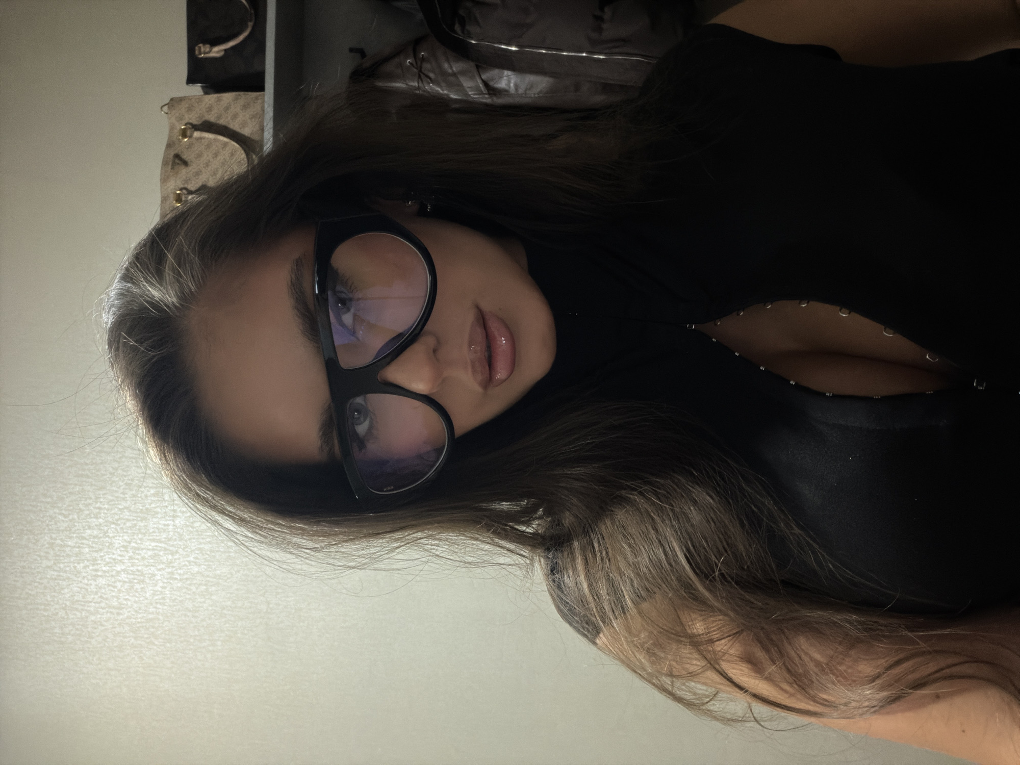 A woman with long brown hair and wearing large, black-framed glasses is facing the camera. She is wearing a black top with a low neckline. The background appears to be a room with a light-colored wall to the left and darker items, possibly bags, behind her head. The image does not contain enough information to determine the location in Krasnoyarsk, Russia.