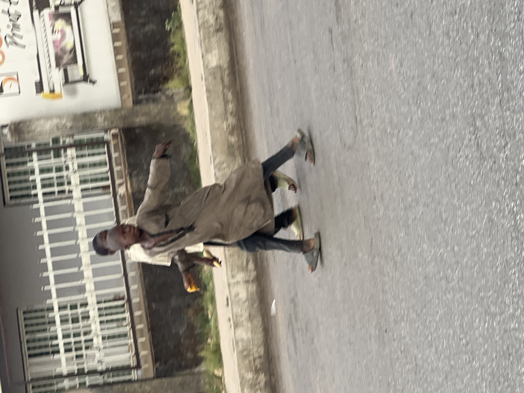 A person is walking on a street in what is presumed to be Jos, Nigeria. The person is wearing a tan coat and dark pants, and appears to be carrying a yellow container in their left hand. The street is paved and there is a sidewalk to the left of the person, along with a building and some green vegetation. The photo appears slightly blurry.