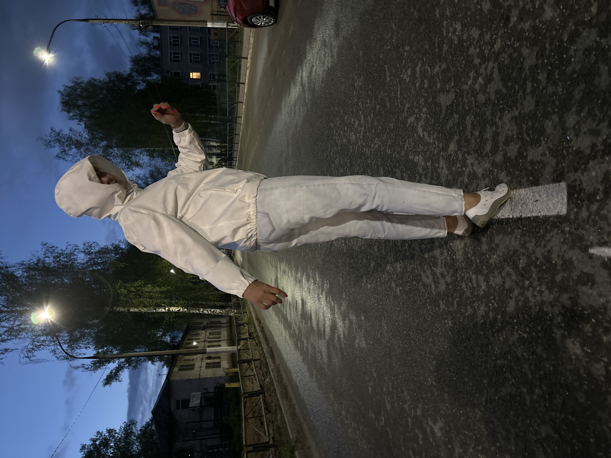 A person in a white hooded jacket and white pants lies on a wet asphalt road. The person's face is obscured by the hood. They are wearing white shoes and their fingernails are painted red. The road has a white line marking on one side. In the background are buildings, trees, and streetlights. It is dusk. The location is in the city of Chaikovsky, Russia.