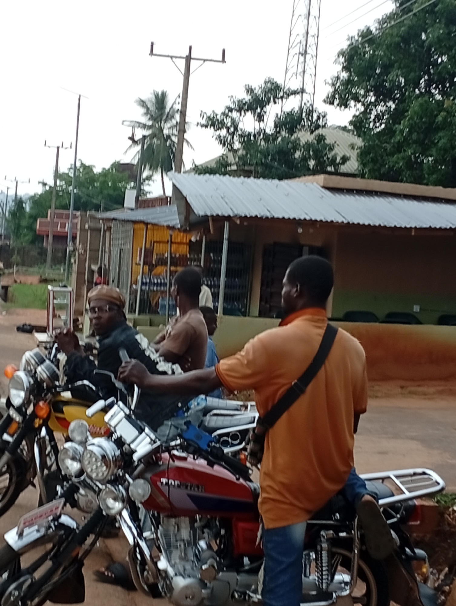 In Nsugbe, Nigeria, a group of individuals are gathered around motorcycles on a street. One man, wearing a brown headwrap and glasses, sits on a yellow and black motorcycle, looking towards the camera. Another man in an orange shirt and blue jeans stands with a black strap across his chest, facing away from the camera. Several other people are visible in the background, some of whom appear to be behind a storefront. The environment includes a street, a building with a corrugated metal roof, and utility poles in the distance. The lighting suggests it is daytime, possibly overcast or late afternoon.
