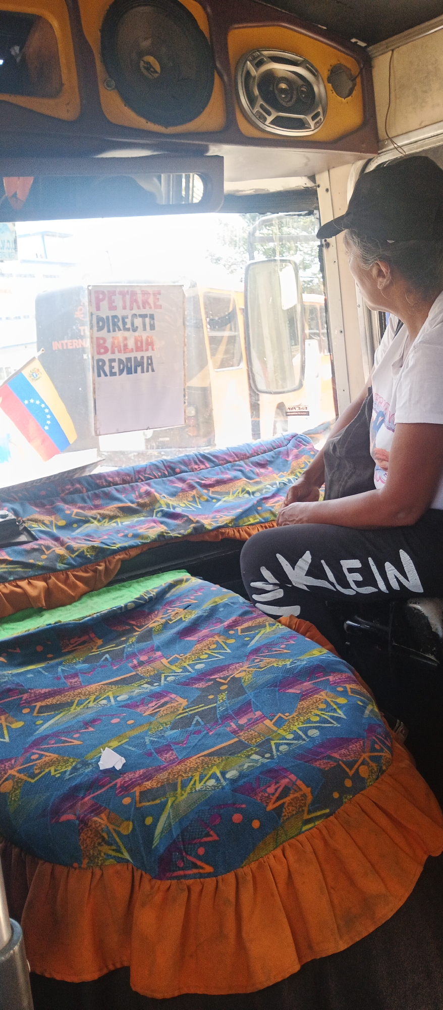 A medium shot shows a woman with dark hair and a black baseball cap sitting in a bus, looking out the window. She wears a white t-shirt and black leggings with "KLEIN" printed on them in white. The bus interior features two colorful, ruffled mattress-like surfaces in the foreground. A sign with red and blue lettering is visible in the window, reading "PETARE DIRECTI BALAA REDIMA" above the Venezuelan flag. The scene is illuminated by daylight filtering through the window.