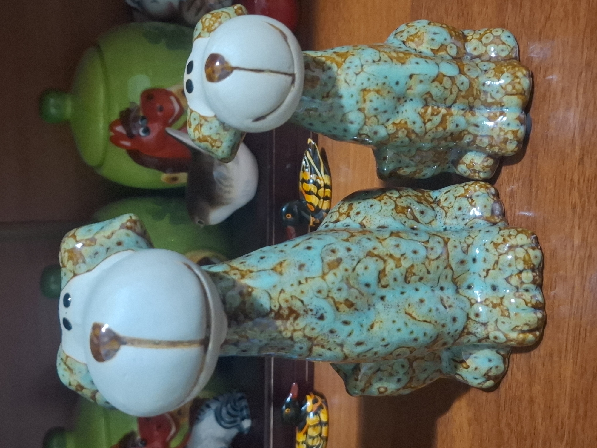 The image shows two ceramic dog figurines with teal and brown speckled glaze, positioned on a wooden surface. The figurines are facing forward, with their heads tilted slightly. In the background, there are other decorative items, including green ceramic jars and what appears to be a horse figurine. There is also a small, decorative duck figurine in the foreground, to the left of the larger dog. The lighting suggests an indoor setting, possibly a shelf or display area. There is no discernible information about the city of Almaty, Kazakhstan, or any specific location within it.