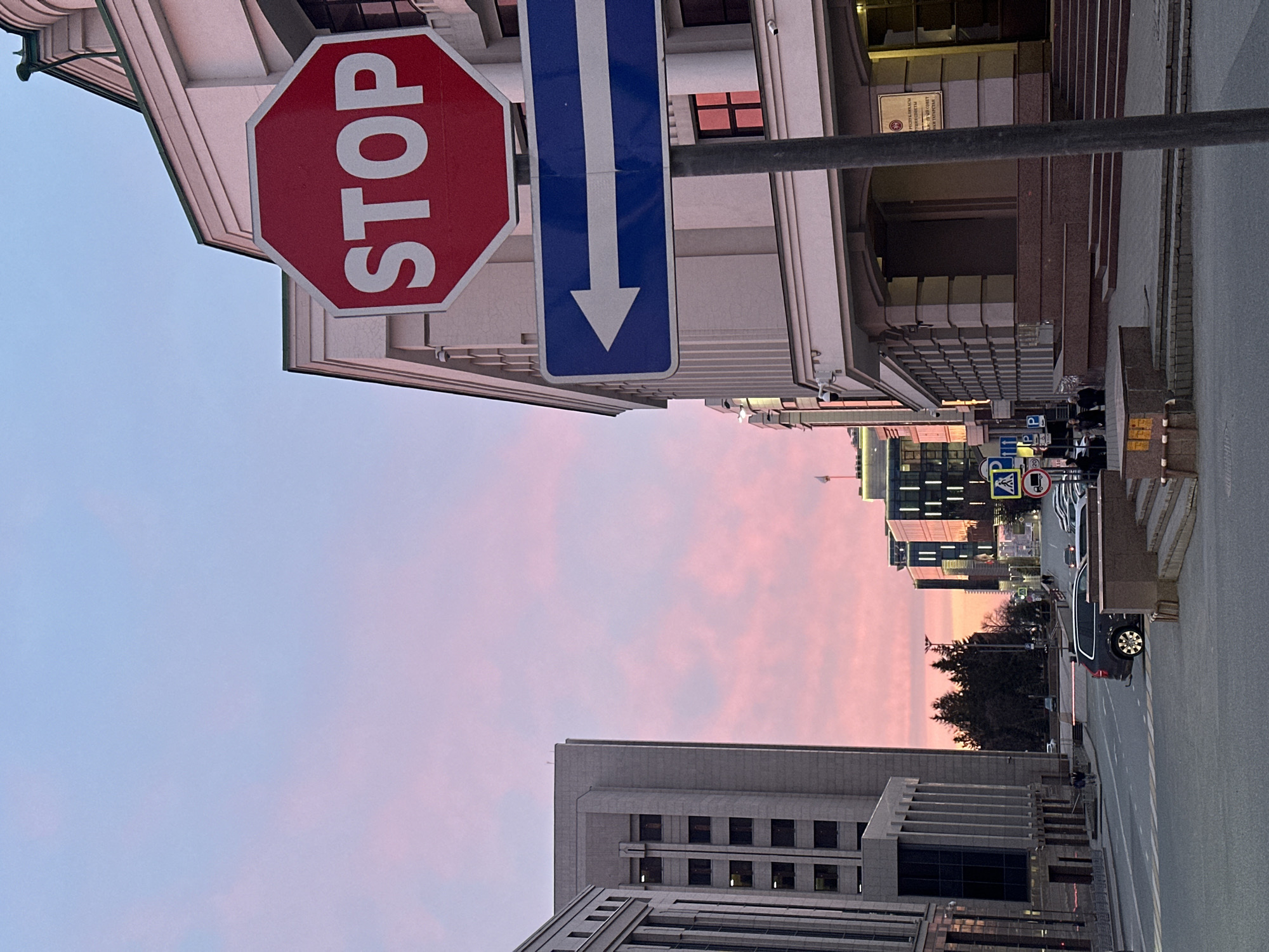 The image shows a street scene in Kazan, Russia, at dusk. The sky is a mix of blue and pink. A stop sign and a blue directional sign hang above the street. Tall buildings line both sides of the street, with some under construction. Vehicles, including a black SUV, are seen on the right side of the street. Several people are visible on the sidewalk. The environment suggests an urban setting.