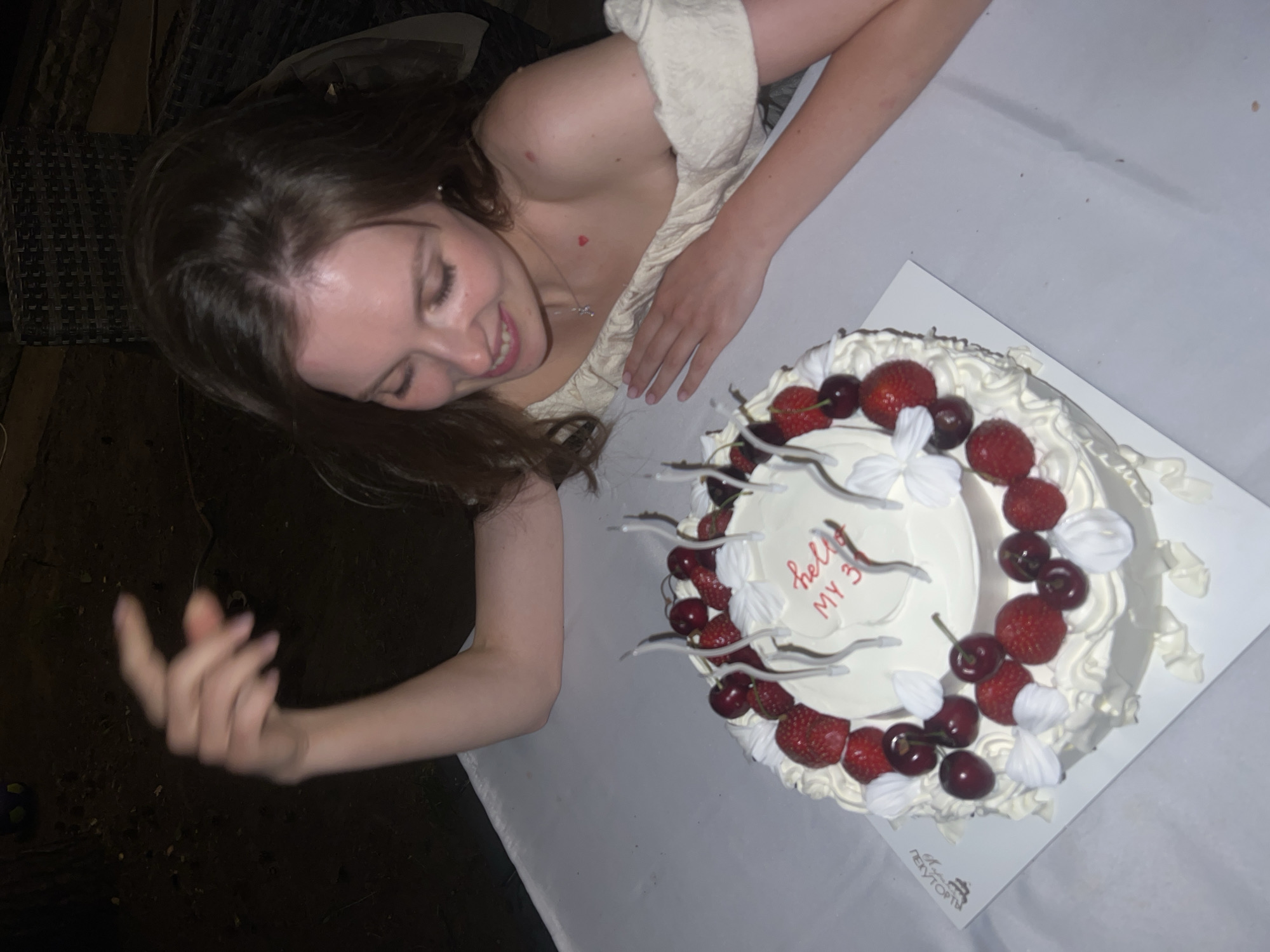 A young woman with long brown hair and fair skin is leaning over a white cake decorated with strawberries and cherries. The cake has "hello MY 3" written in red icing and several unlit candles. The woman is wearing a cream-colored off-the-shoulder top and a necklace with a small charm. Her right arm is extended and her hand is in a blurred motion above the cake. The background is dark and appears to be an outdoor setting with some wicker furniture visible. The overall scene suggests a birthday celebration. There is no information in the image that indicates the location is in Tomilino, Russia.