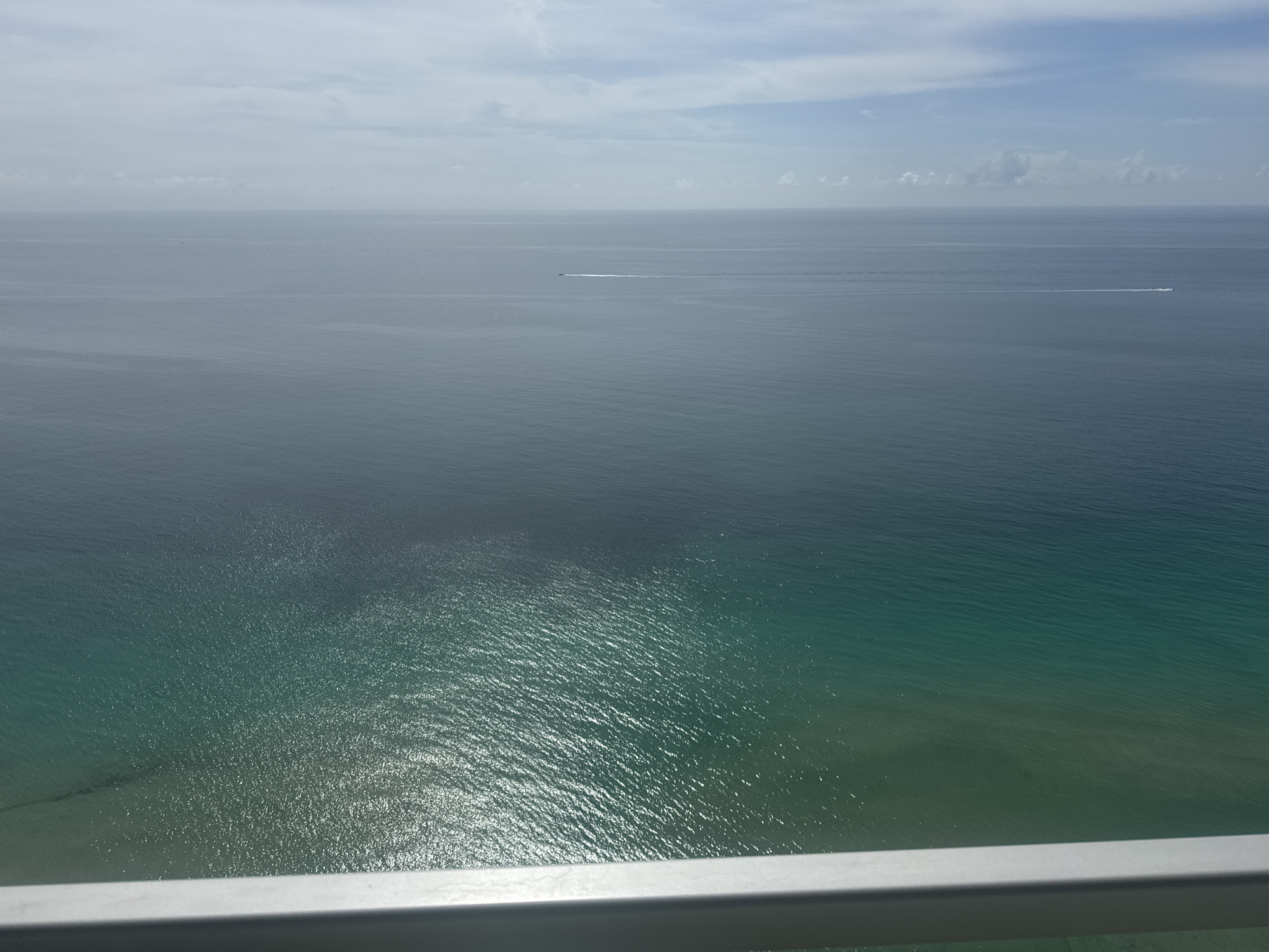 The image shows a wide expanse of the ocean under a partly cloudy sky. The water transitions in color from a dark blue-grey at the horizon to a lighter, more turquoise hue in the foreground. Sunlight reflects off the water, creating shimmering patterns. A small boat can be seen leaving a trail in the distance. The bottom edge of the image shows a metallic railing.