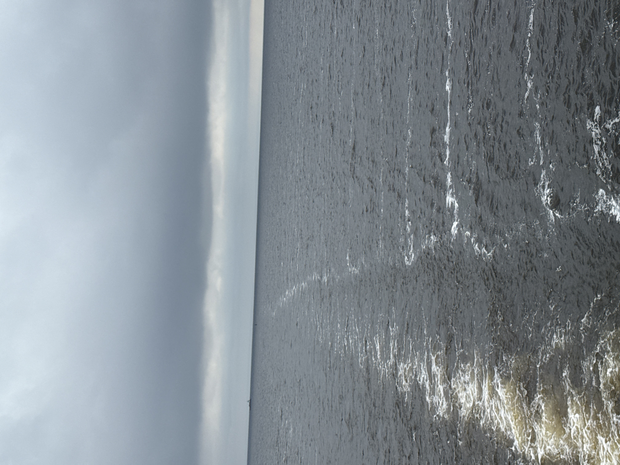 This is a vertical, eye-level, medium shot of a body of water under a cloudy sky. The water is dark and appears to be a lake or sea. There is a distinct wake trailing from the bottom left corner of the frame towards the middle distance, indicating a vessel has recently passed through. The wake is frothy and white in the foreground, becoming less pronounced as it moves away. In the far distance, on the horizon, a small, dark object that could be a boat or buoy is visible. The sky is overcast with a thick layer of gray clouds that dominate the upper portion of the image, obscuring any sunlight. The horizon line is sharp and defined, separating the water from the sky. The location is Laishevo, Russia.