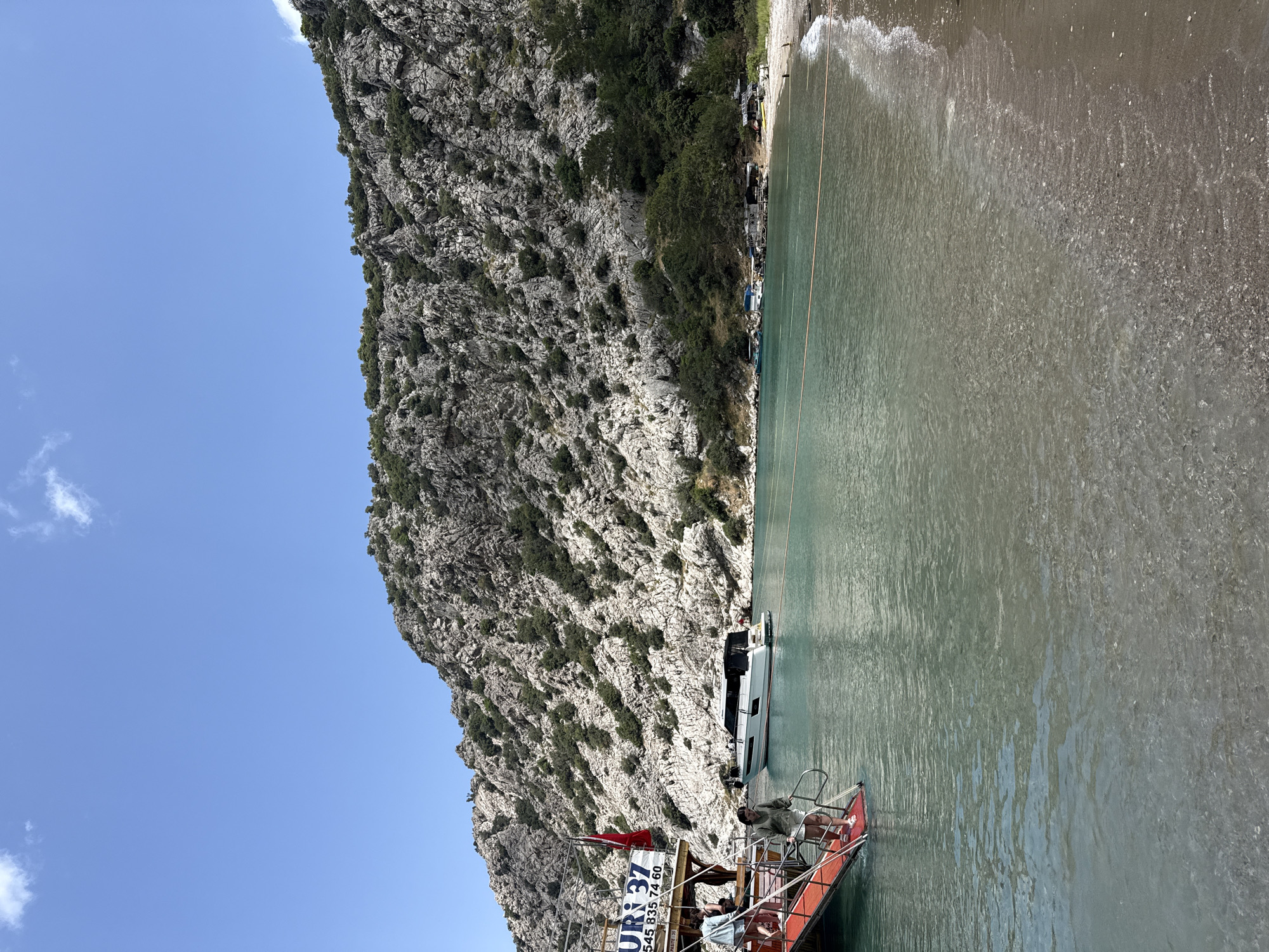 The image shows a scenic view of a rocky mountain next to a body of water, likely a bay or cove, in Kumluca, Turkey. The mountain face is predominantly grey and rocky with sparse vegetation clinging to its surface. A clear blue sky is visible above the mountain. Adjacent to the mountain is a body of clear water. A sandy beach borders the water. Several boats, including a tour boat, are docked along the shore. Some people are on the tour boat.