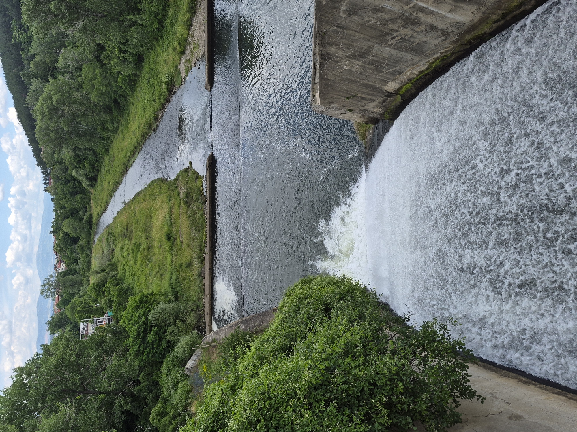 A high-angle, vertical, full shot captures a wide, powerful waterfall cascading down a concrete dam. The turbulent white water fills the foreground, frothing and churning as it descends. On the left, lush green foliage spills over the edge of the dam, contrasting with the gray concrete structure. The dam itself is reinforced with concrete walls, showing signs of age with cracks and mossy patches.
Beyond the dam, a river flows into the distance, its surface rippling. Further upstream, the river narrows and meanders through a verdant landscape of grassy banks and dense trees. The river appears to be contained within a channel with concrete embankments in places. In the far background, a town with red-tiled roofs is nestled amongst hills covered in trees, and distant mountains form the horizon under a sky with scattered white clouds. The scene is a natural landscape enhanced by man-made structures, a dam controlling the flow of a river within a semi-rural or suburban environment. There are no people or animals visible in the frame.