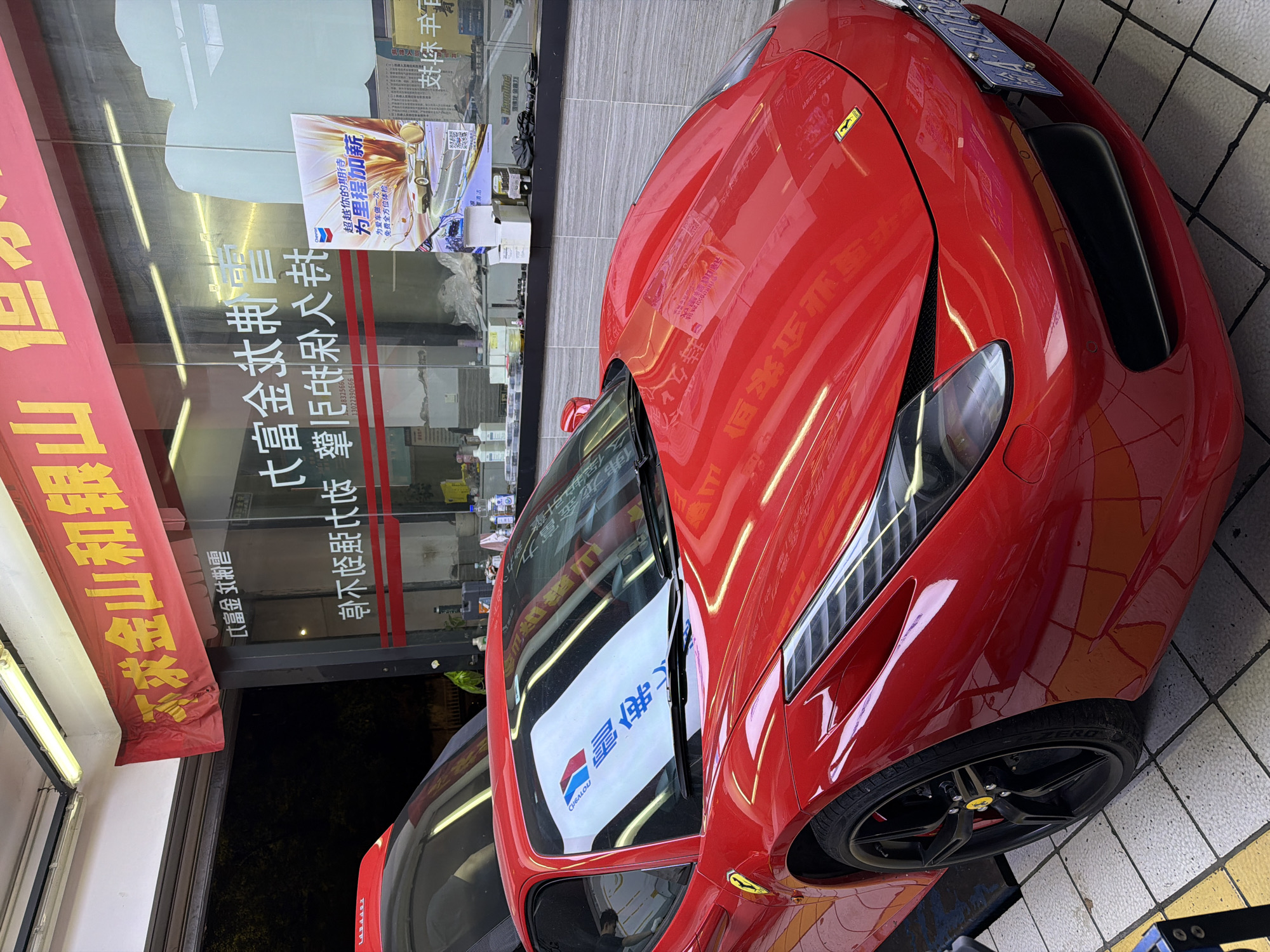 This media file, captured in Chongqing High-tech Zone, China, depicts a vibrant red Ferrari sports car parked outside a building, likely a service center or store, during the evening. The car, with its distinctive headlights, front grilles, and black multi-spoke wheels adorned with yellow Ferrari emblems, is the central subject. Its glossy surface reflects the surrounding artificial lighting and parts of the storefront. A Chinese license plate is visible on the front of the car, and "ZERO" is visible on the Pirelli tires.
The background features a glass storefront with multiple reflections and posters. A red awning above the storefront displays Chinese characters, including "富金期震" and "不求金山和银山". Reflected on the glass are additional Chinese characters like "大富金典" and phone numbers "132566" and "13022390666". A prominent advertisement on the window features the Chevron logo and text that translates to "Beyond your expectations," "Add new for mileage," and "Give your beloved car a free comprehensive check-up." It also promotes "Long-lasting, long-term engine power" and mentions "Havoline Jin Fuli" and "Chevron Jin Fuli" products. The Chevron logo is also reflected on the car's windshield.
The scene is illuminated by artificial lights, suggesting it is dark outside. The car rests on a light-colored tiled floor with a yellow demarcation, indicating a commercial or service area in an urban environment. No people are clearly visible, but a faint reflection of a person can be seen on the car's windshield.