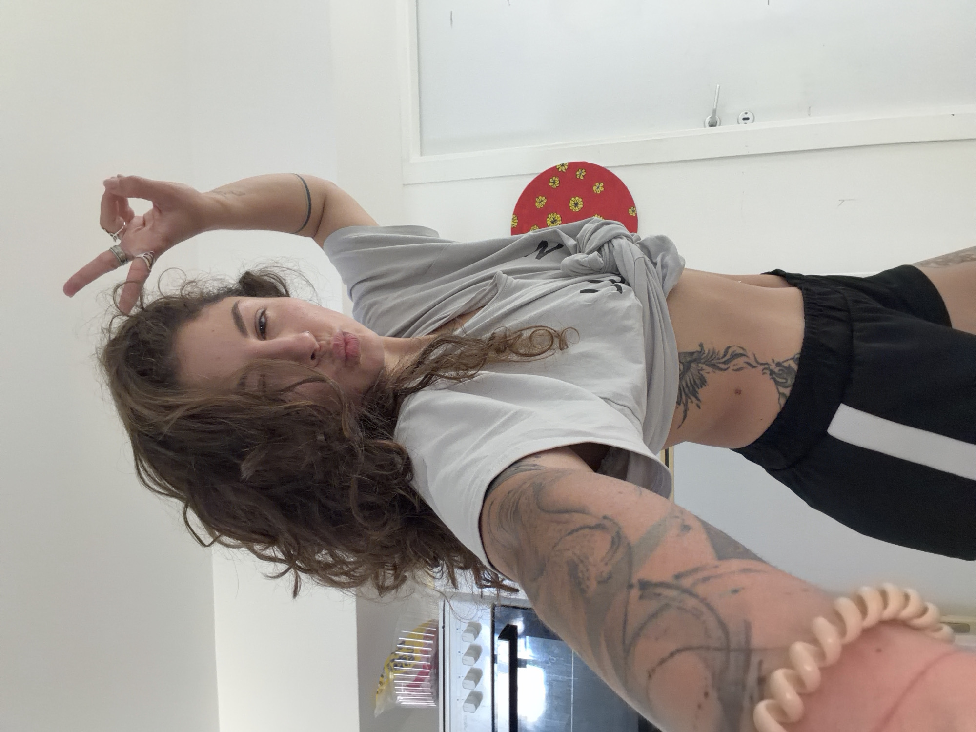 A young person, likely female, with long, wavy dark brown hair, poses for a selfie indoors in Kiryat Bialik, Israel. The image is taken from a low angle, rotated slightly clockwise, giving it a dynamic feel. The subject is looking directly at the camera with a slight pout.
She is wearing a light grey short-sleeved t-shirt, tied in a knot at the front to expose her midriff, and black athletic shorts with a white stripe on the side. Extensive dark tattoos cover both her arms and a portion of her left torso/rib cage, featuring intricate, possibly abstract or floral designs. On her left hand, which is held up in a stylized gesture with bent fingers, several rings are visible. A light-colored, spiral-coil wristband is worn on her right wrist.
The background consists primarily of bright white walls. Above her, a red circular object adorned with yellow floral patterns is mounted on the wall, possibly a decorative item or part of a skateboard. To her right, a white counter or shelf is visible with what appears to be a microwave or oven on it. The lighting is bright and even, suggesting it is daytime. The overall mood conveyed is playful and confident. No clearly legible text is visible on the clothing or in the background.