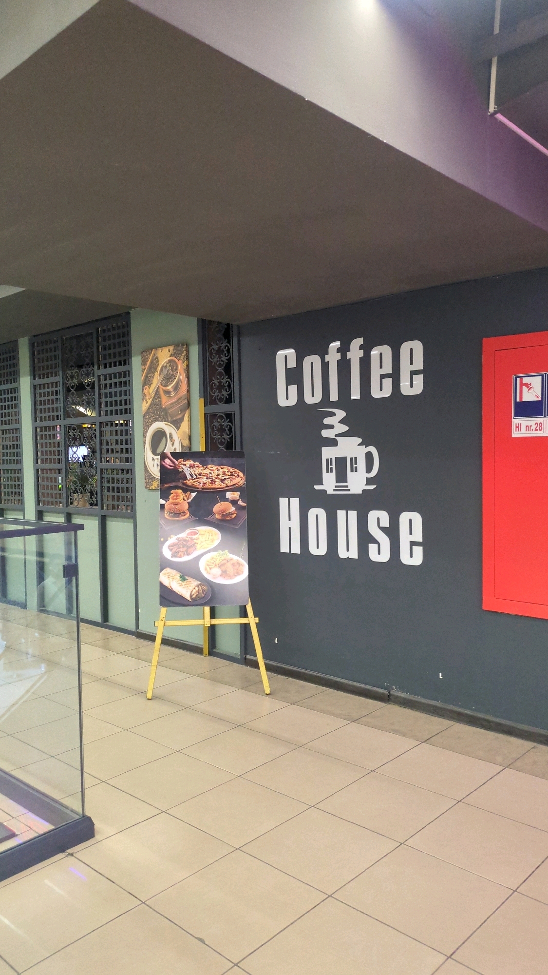 The image shows a storefront with a large sign that reads "Coffee House". The sign is white and in a bold, sans-serif font. The background of the sign is dark gray. To the left of the sign is a display easel with a menu of food items, including pizza, burgers, and wraps. The easel is yellow. To the right of the sign is a red fire extinguisher cabinet with a sign that reads "HI nr. 28". The storefront appears to be in an indoor mall or shopping center, with tiled flooring and a gray ceiling. The overall impression is that of a modern, casual dining establishment.