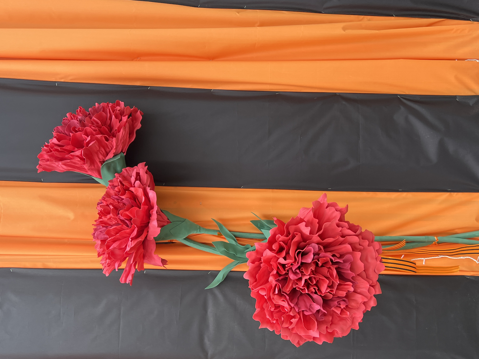 The image shows an arrangement of three large, artificial red carnations laid against a backdrop of alternating orange and black stripes. The arrangement appears to be indoors. The carnations are fully bloomed with ruffled petals and green stems. The striped background suggests a commemorative or decorative purpose, possibly linked to Victory Day celebrations in Russia. The location Chaikovsky, Russia, is specified, indicating this may be a local decoration.