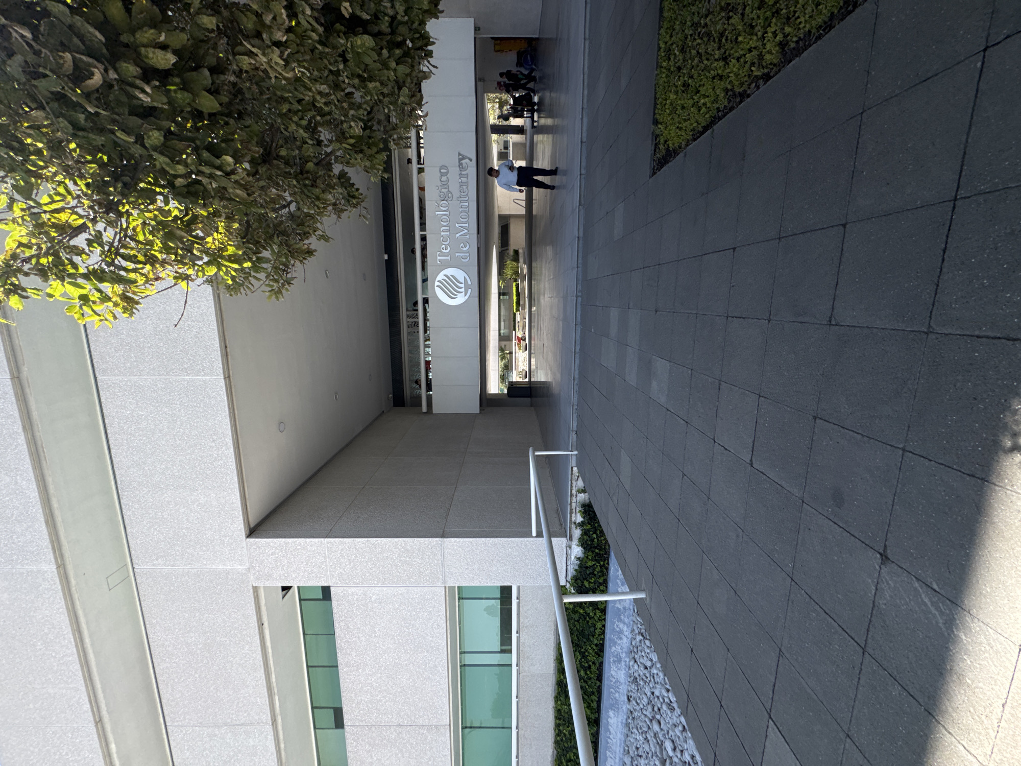 The image captures an outdoor scene on a bright, sunny day, likely within the campus of Tecnológico de Monterrey in Heroica Puebla de Zaragoza, Mexico. The central focus is a modern building entrance with a prominent sign displaying "Tecnológico de Monterrey" and its distinctive logo, featuring stylized wavy lines.
A man dressed in a light blue shirt and dark trousers is walking on the paved pathway, appearing to be exiting the building. In the background, near the entrance, other individuals are visible, suggesting general foot traffic. The building itself boasts a contemporary architectural style with a light-colored, possibly stone or concrete, facade and large, reflective glass windows on the lower levels. An overhead awning provides shade for the entrance area.
To the left of the frame, a large tree with abundant green foliage is brightly illuminated by the sun, casting dappled shadows. The ground features dark grey square paving stones, forming a clean walkway. To the right, a landscaped area includes low green shrubbery and decorative white pebbles, bordered by metal railings. The strong sunlight creates clear shadows, indicating a midday setting. The scene suggests a well-maintained, functional, and active educational or institutional environment.