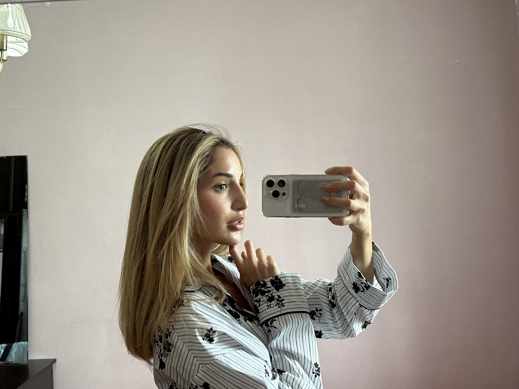 A young woman with blonde hair is taking a selfie in a room with pink walls. She is wearing a black and white striped pajama set with black floral details on the cuffs and collar. She is holding a smartphone in her right hand, which is reflecting a lamp in the upper left corner of the image. Her left hand is resting on her chin. The background is a plain pink wall, with a dark object, possibly a dresser or wardrobe, visible on the left side of the frame. The scene appears to be an indoor domestic setting, likely a bedroom. There is no information to suggest the location is in Moscow, Russia.