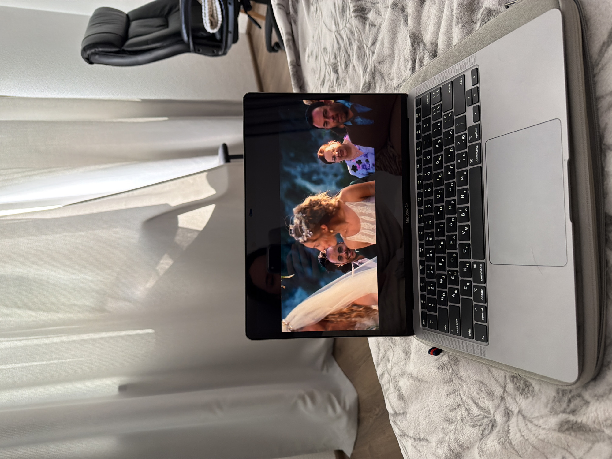 An overhead medium shot shows a MacBook Air laptop computer resting on a grey, plush blanket on the floor. The laptop screen displays a video of a wedding ceremony. In the foreground of the screen, a woman in a white dress and veil is partially visible. To her right, a woman in a floral dress is looking to the left. To the right of the woman in the floral dress, a man in a dark jacket and blue shirt is looking forward. In the background of the screen, a person is reflected in the laptop screen, holding a phone. To the left of the laptop, white sheer curtains are visible, backlit by sunlight. In the background to the right, a black leather office chair with a knitted blanket draped over it is positioned against a white wall. The flooring visible in the background is wood. The scene appears to be in an indoor domestic setting.