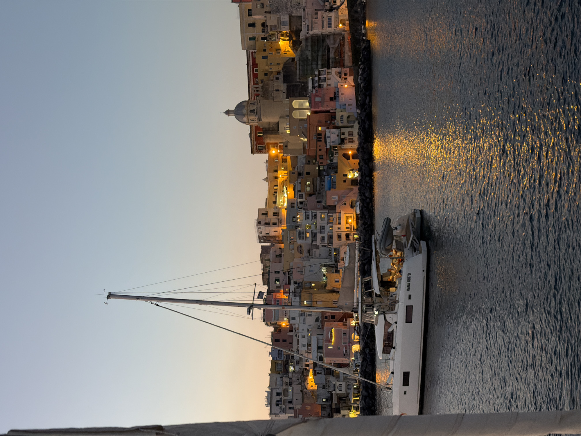 The image shows a vertical view of a boat in the harbor of Procida, Italy at dusk or dawn. The port of Procida, with its colorful buildings, is visible along the harbor. A white sailboat is docked near the buildings, and the water reflects the lights from the town. The sky is a gradient of light blue to orange.