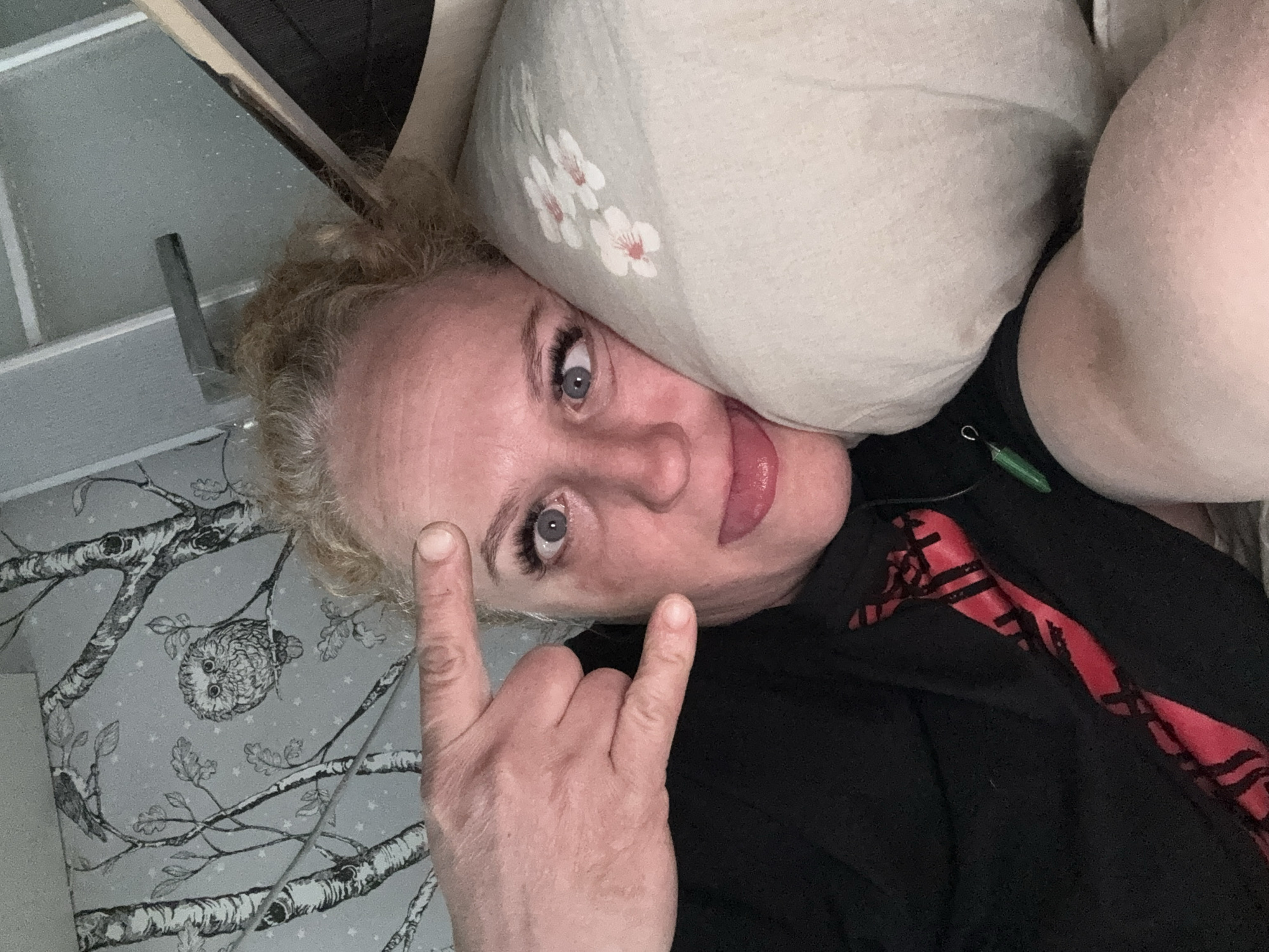 A woman with curly blonde hair, blue eyes, and pink lipstick is lying down, propped up by a pillow with a floral pattern. She is wearing a black shirt with a red design. She is making a rock-and-roll hand gesture. The background features wallpaper with a tree and owl pattern. The image was taken indoors. There is no evidence to confirm the location is Chaikovsky, Russia.