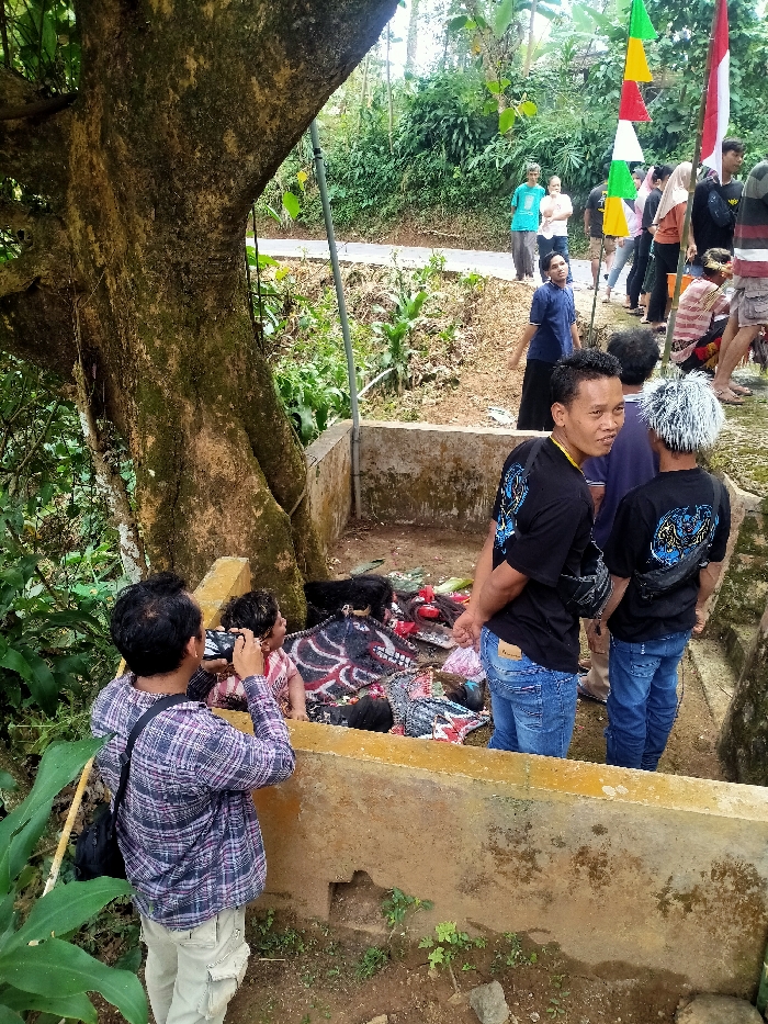 A public gathering is depicted in an outdoor, semi-rural environment in Ngargoyoso, Indonesia. In the foreground, a male individual wearing a plaid shirt and light-colored cargo pants is seen from behind, holding a smartphone to photograph the contents of a U-shaped concrete enclosure. Inside this enclosure, located at the base of a large, moss-covered tree trunk, various objects are arranged on the ground. These objects include fabric items, possibly masks or effigies, one displaying a red and black pattern, another with a white and black design, along with dark, possibly fibrous materials.
To the right of the enclosure, two male subjects stand, observed from their backs and sides. Both wear black t-shirts featuring a blue and black emblem on the back and blue jeans. One has spiky, light-colored hair, while the other has dark hair and is looking towards the left foreground. Both wear black fanny packs.
In the background, on an elevated dirt path or road, multiple individuals are gathered, observing the scene. Notable figures include a male in a light blue t-shirt and another in a dark blue shirt. Several triangular flags (green, yellow, red, white) are visible on a pole in the background. The environment is characterized by lush green foliage and the weathered concrete structures of the enclosure.