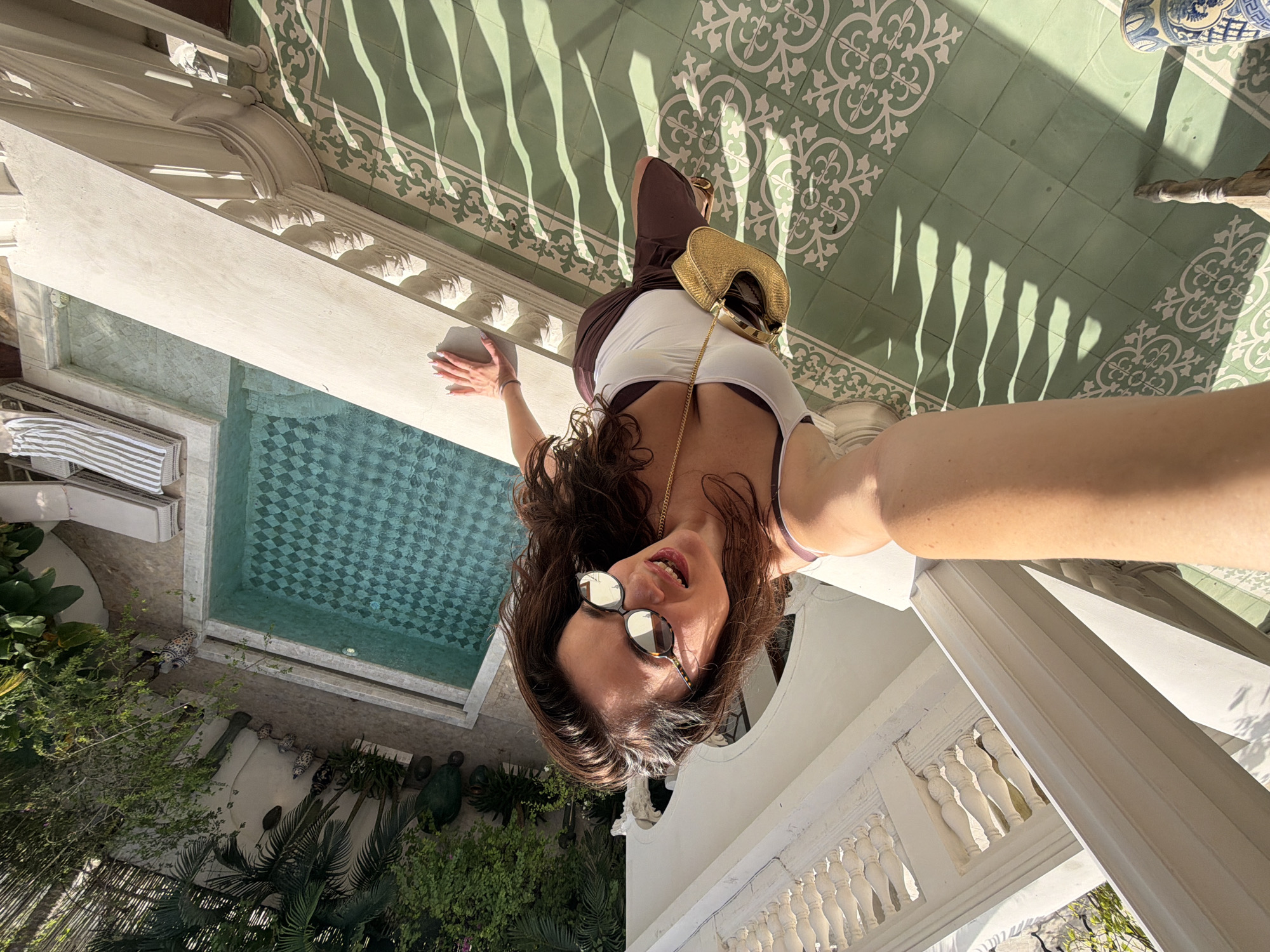 An overhead, inverted selfie captures a woman smiling and looking directly at the camera from a balcony or elevated walkway. She has dark, wavy hair, wears sunglasses, a white top over a dark bra, and a dark lower garment, accessorized with a small, gold-colored woven bag with a chain strap. Her left hand rests on a decorative white balustrade.
The surface beneath her feet features distinctive green and white patterned tiles, across which long, wavy shadows are cast, indicative of strong, direct sunlight. Below the balcony, a turquoise swimming pool with a checkered tile bottom is visible. The pool area is surrounded by lush green tropical foliage, including palms and shrubs, along with white lounge chairs and various decorative ceramic or stone objects. The architecture is characterized by white ornate pillars, balustrades, and intricate carvings, suggesting a luxurious or traditional design.
The scene is brightly lit, indicating a sunny day, likely midday or early afternoon, creating a vibrant and relaxed atmosphere. The setting, with its tropical greenery, pool, and architectural style, is consistent with a resort or villa in a warm climate such as North Kuta, Indonesia. No visible text is present in the image.