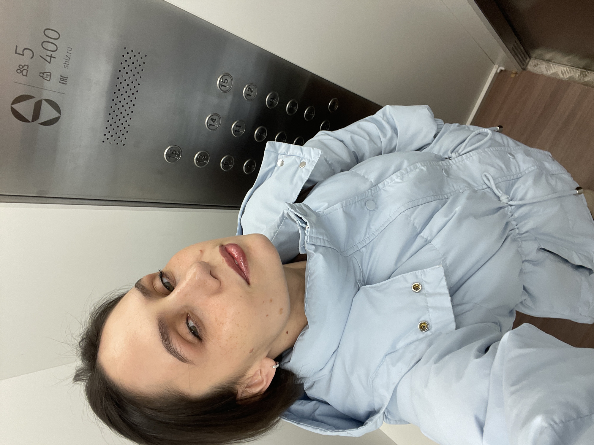 A woman stands inside an elevator. The elevator control panel is visible, with buttons labeled 5, 16, 14, 15, 13, 12, 11, 10, 9, 8, 7, 6, 400. The panel also includes symbols and the text "shlz.ru." The elevator's floor appears to be made of wood and is in the city of Izhevsk, Russia. The woman is wearing a light blue padded jacket. She is looking directly at the camera. Her expression is neutral.