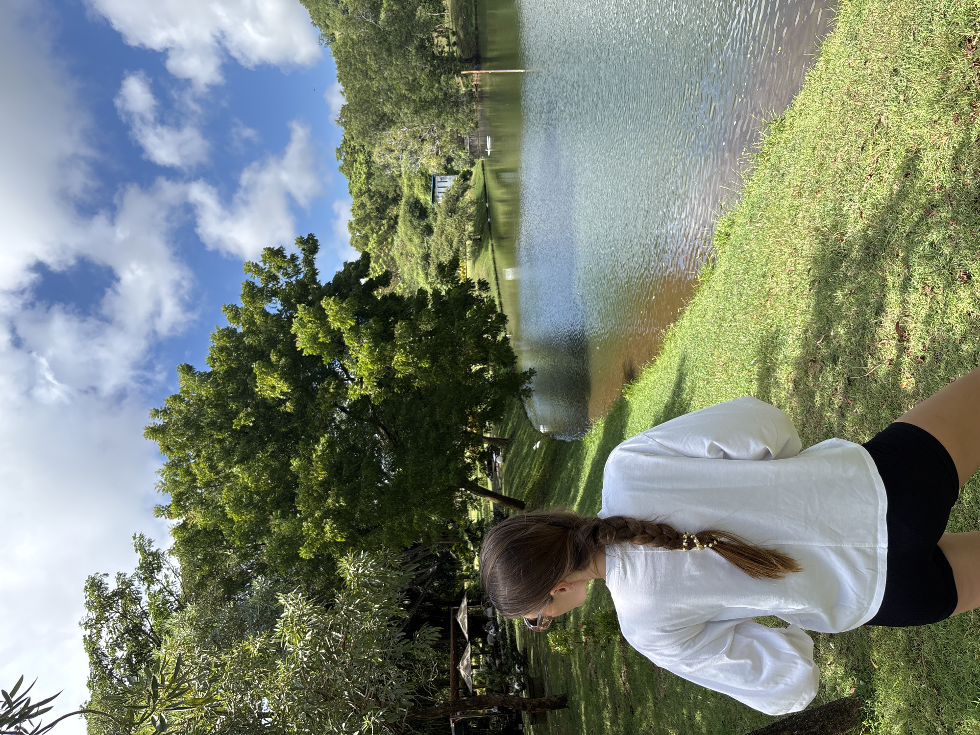 A woman stands near a lake or body of water in Kuta Selatan, Indonesia. The lake is surrounded by trees and green vegetation. The sky is blue with white clouds. The woman has her back to the camera, and she is wearing a white shirt and black shorts. She has a braid in her hair.