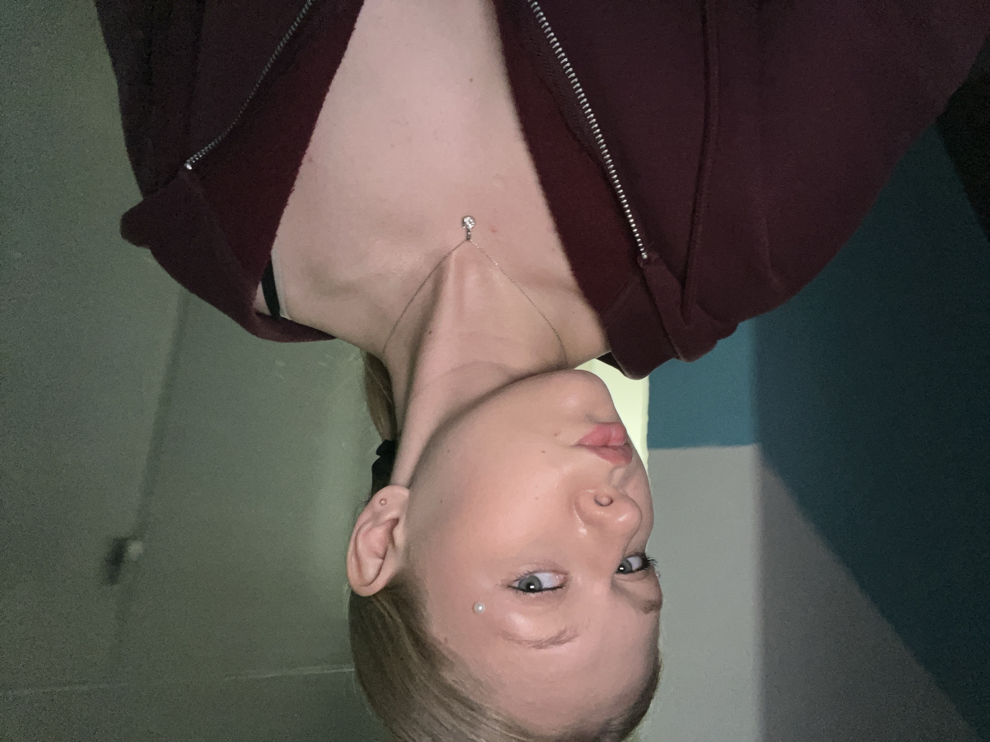 A young woman with blonde hair pulled back in a ponytail and blue eyes is looking at the camera. She is wearing a dark red zip-up hoodie and a silver necklace with a clear pendant. She has a small piercing on her cheekbone and another on her earlobe. The background is a dimly lit room with walls that appear to be painted in contrasting shades of green and blue. A light blue stripe is visible on the left side of the background. The lighting is dim, casting shadows on the wall behind her. The image appears to be a selfie taken in a residential building, likely an apartment or hallway, in the city of Perm, Russia.
