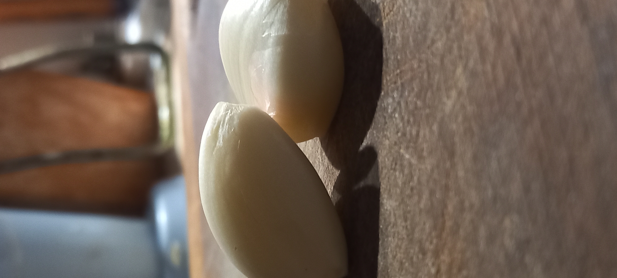 The image presents a close-up, horizontally oriented shot of two peeled garlic cloves resting on a textured wooden surface. The garlic cloves, light cream to off-white in color, are in the foreground and sharp focus, exhibiting natural surface textures and minor irregularities. One clove is positioned slightly higher and behind the other.
Distinct shadows are cast by the garlic cloves onto the wooden surface, indicating a light source from the upper left, possibly natural light from a window or strong artificial illumination. The wooden surface beneath the garlic displays visible grain patterns and minor imperfections, consistent with a cutting board or kitchen tabletop.
The background is significantly blurred, featuring soft, out-of-focus elements. Warm brown tones are visible in the upper left background, suggesting wooden cabinetry or furniture, while cooler grey tones appear in the lower left, possibly hinting at kitchen appliances or another part of a countertop.
No specific activity is depicted, but the peeled state of the garlic suggests preparation for cooking. There are no people visible, nor is there any discernible text. The scene conveys a simple, domestic setting, likely a kitchen, in Kryvyi Rih, Ukraine.